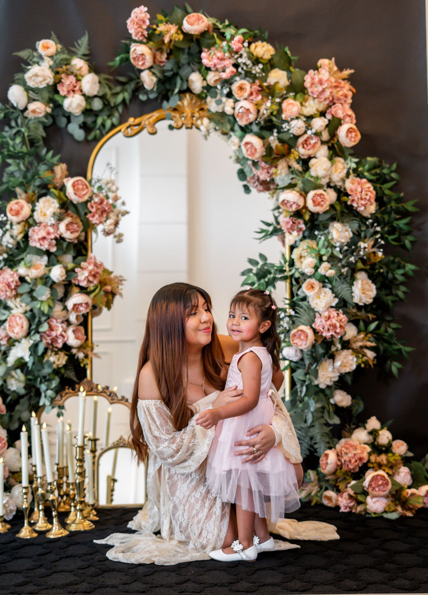 Woman in lace dress poses with toddler girl in pink tutu before elegant floral arch with roses and greenery.