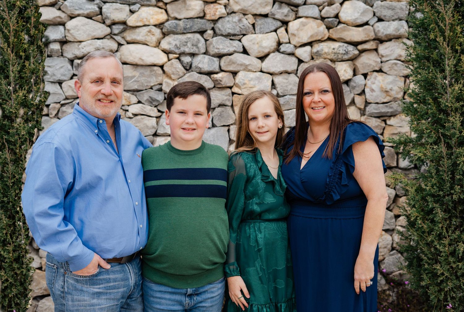 A happy family of four poses in front of a stone wall, dressed in blue and green outfits.