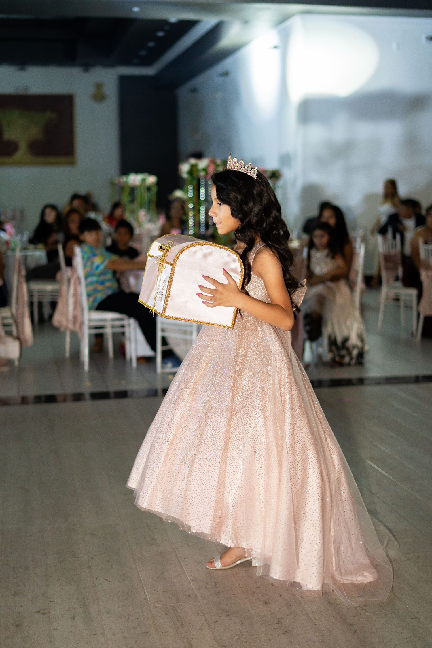 Young girl in pink glitter ball gown and tiara carries decorative box on quinceañera dance floor.