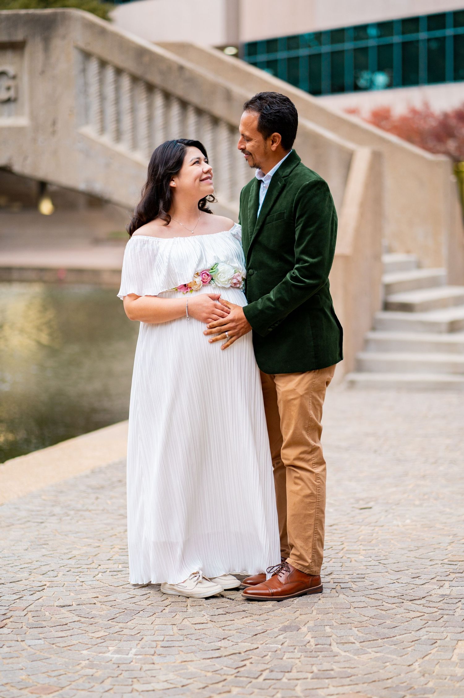 Smiling pregnant couple sharing joyful moment during outdoor maternity photo session by waterway.