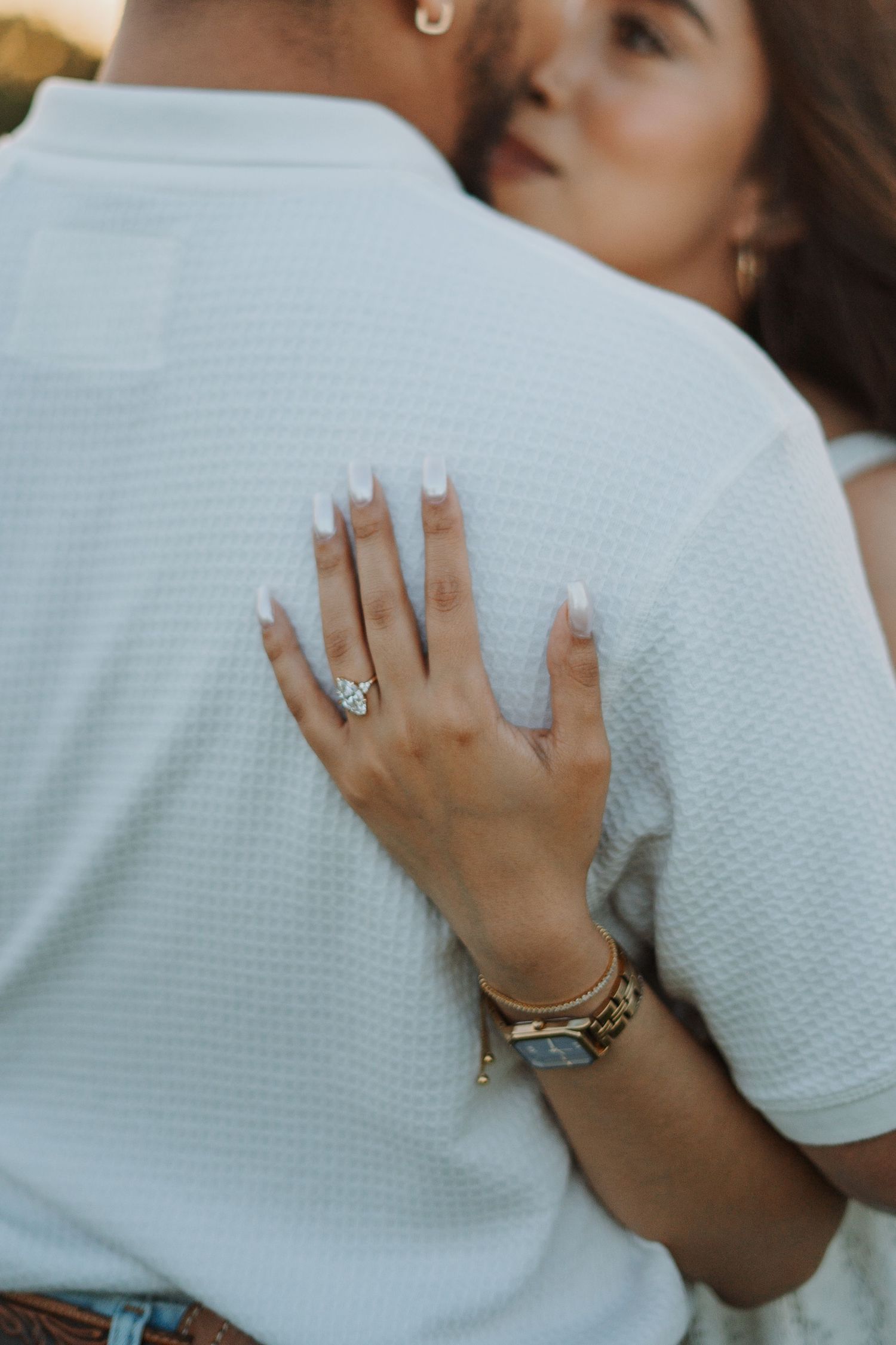 Couple embracing closely, woman's hand on man's back showing diamond engagement ring, gold watch, and bracelets.