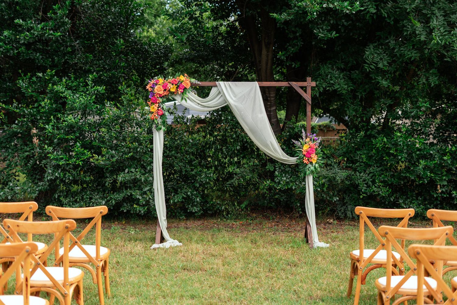 Outdoor wedding ceremony arch with white draping and colorful floral arrangements on green lawn surrounded by lush trees.