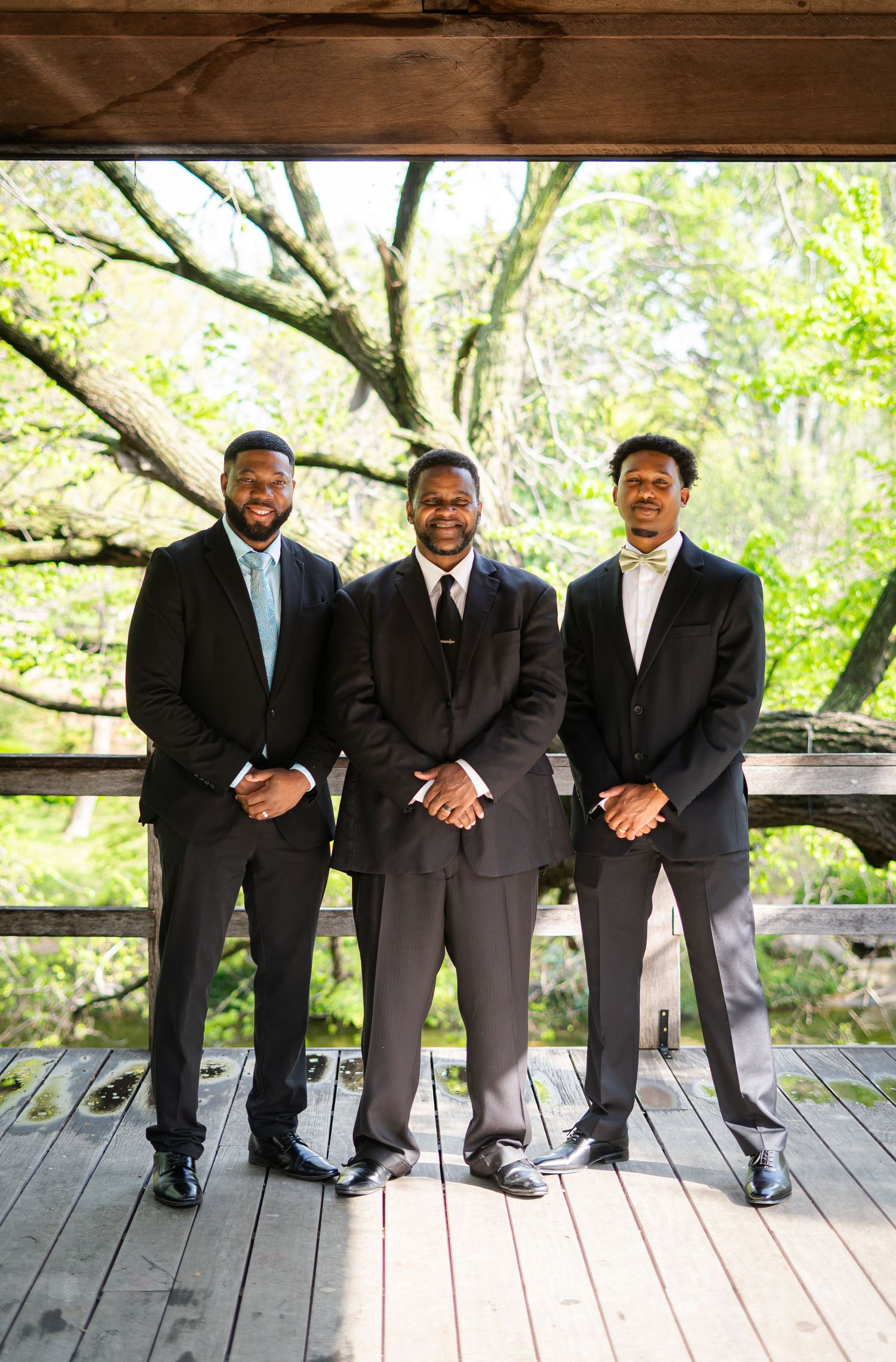 Three men in dark suits pose together on a wooden deck at an outdoor wedding ceremony.