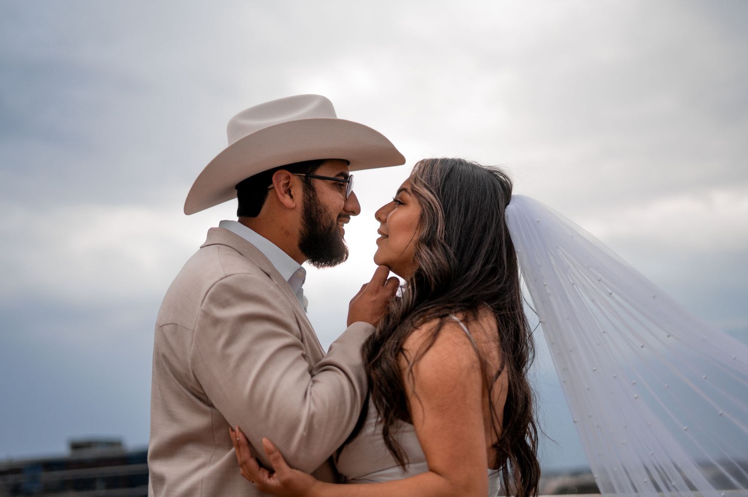 A groom in a white cowboy hat tenderly holds his bride's chin as they gaze at each other under cloudy skies.