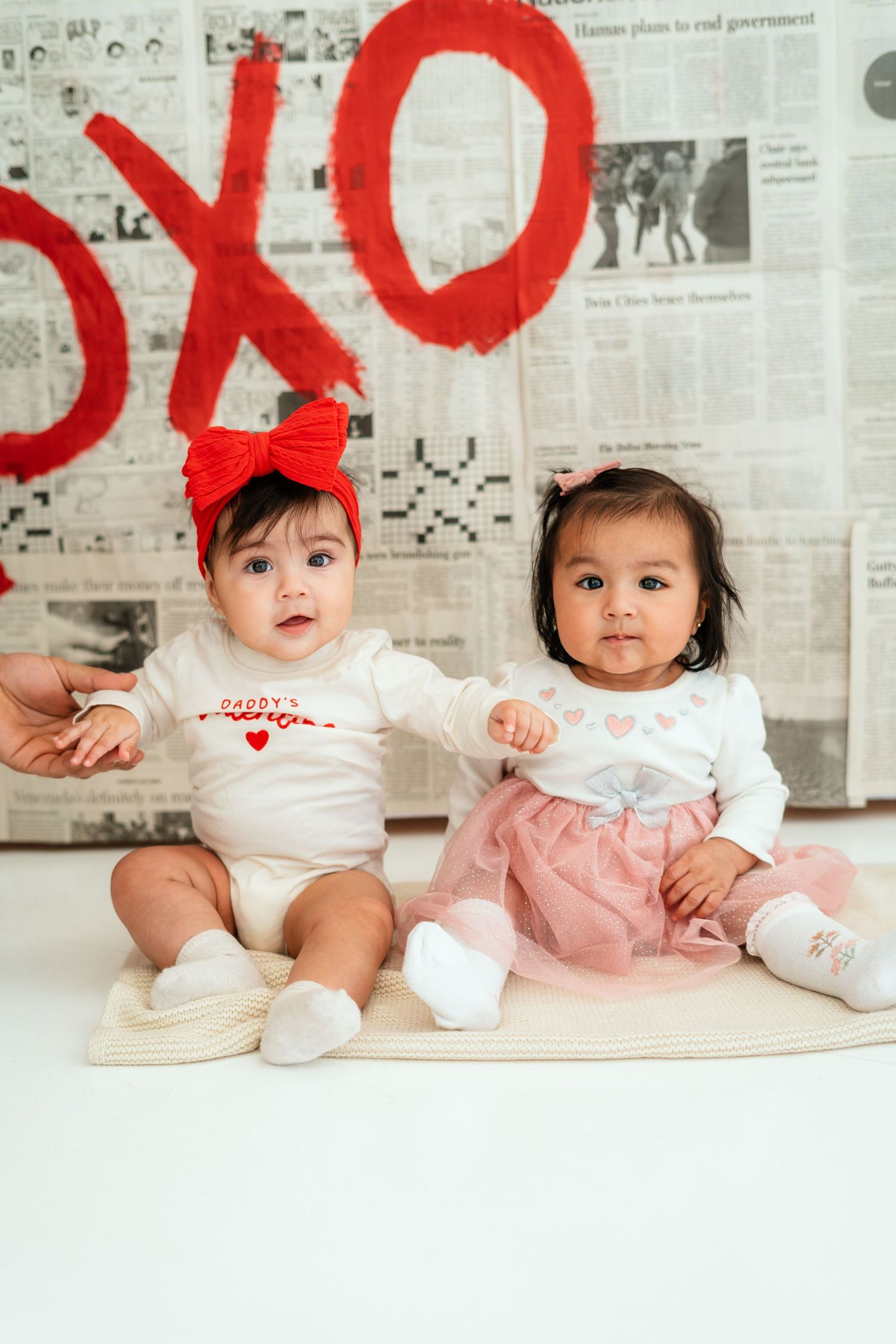 Two cute babies in Valentine's Day outfits sitting before a newspaper XOXO backdrop.