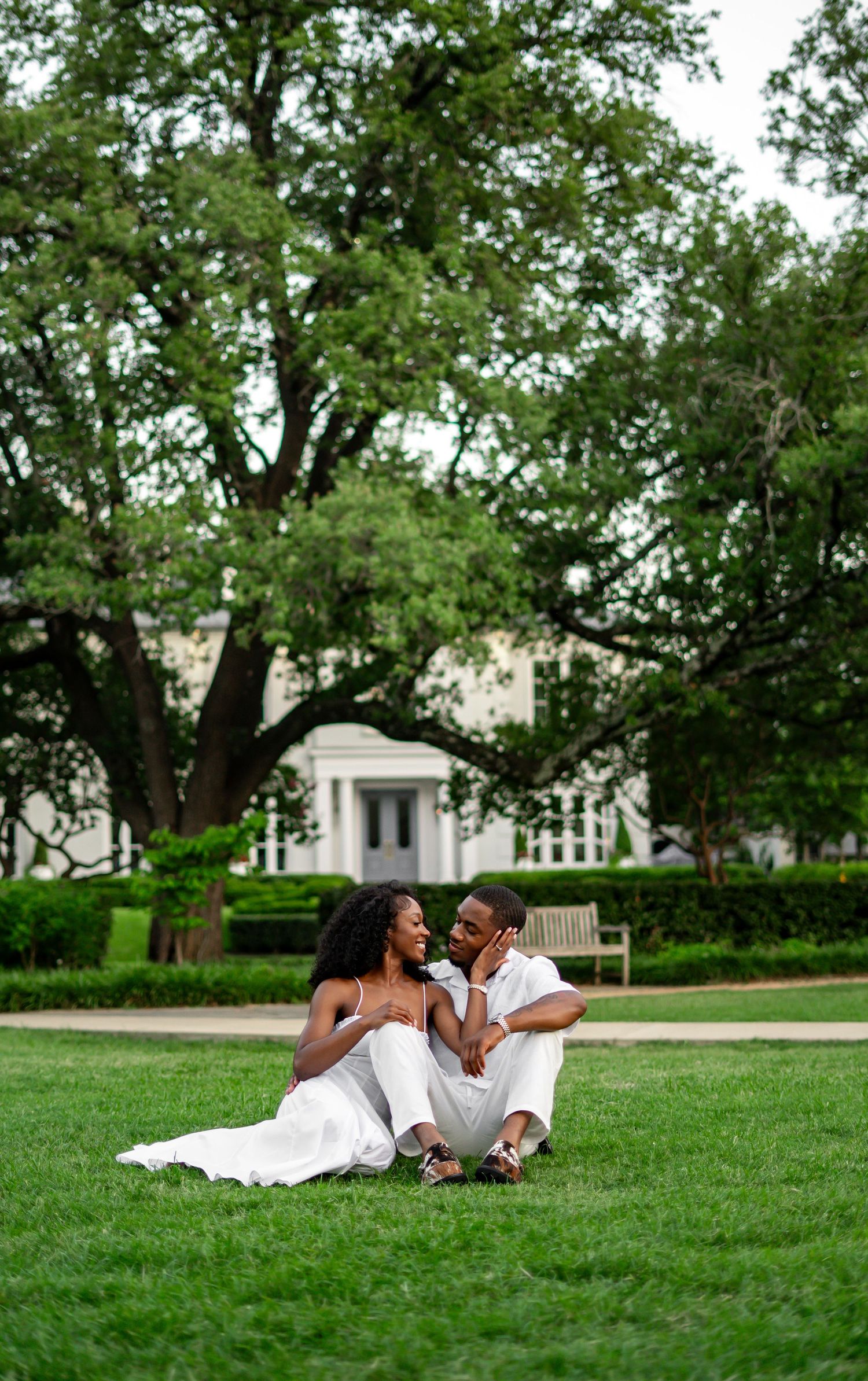 A Black couple in matching white outfits sit intimately on green grass gazing at each other near a white building.