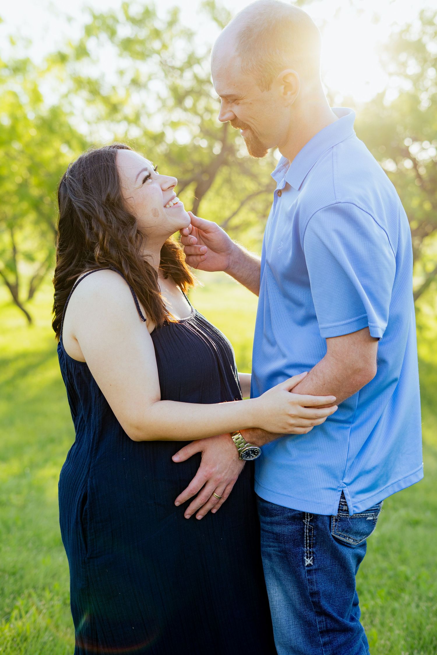 Pregnant couple embracing and smiling at each other in a sunlit green park.