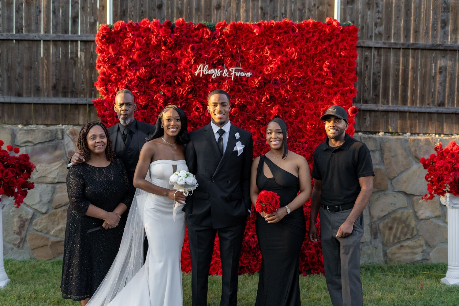 Bride and groom pose with family members in front of red rose flower wall at backyard wedding.