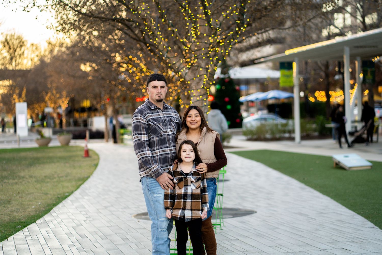 A family of three poses together on a brick pathway surrounded by warm holiday lights and greenery.