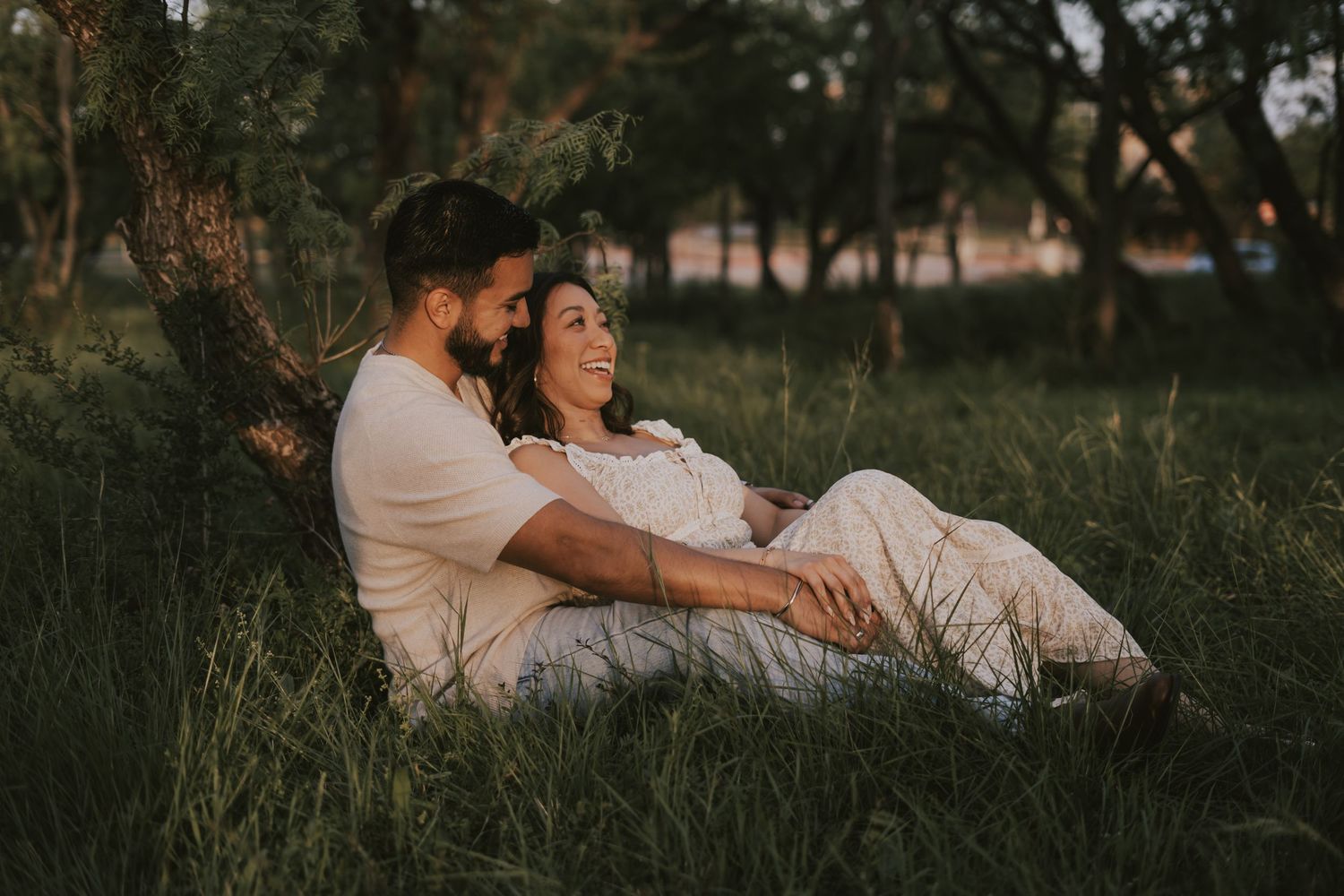 A couple sits together in lush green grass leaning against a tree, sharing a tender romantic moment at golden hour.