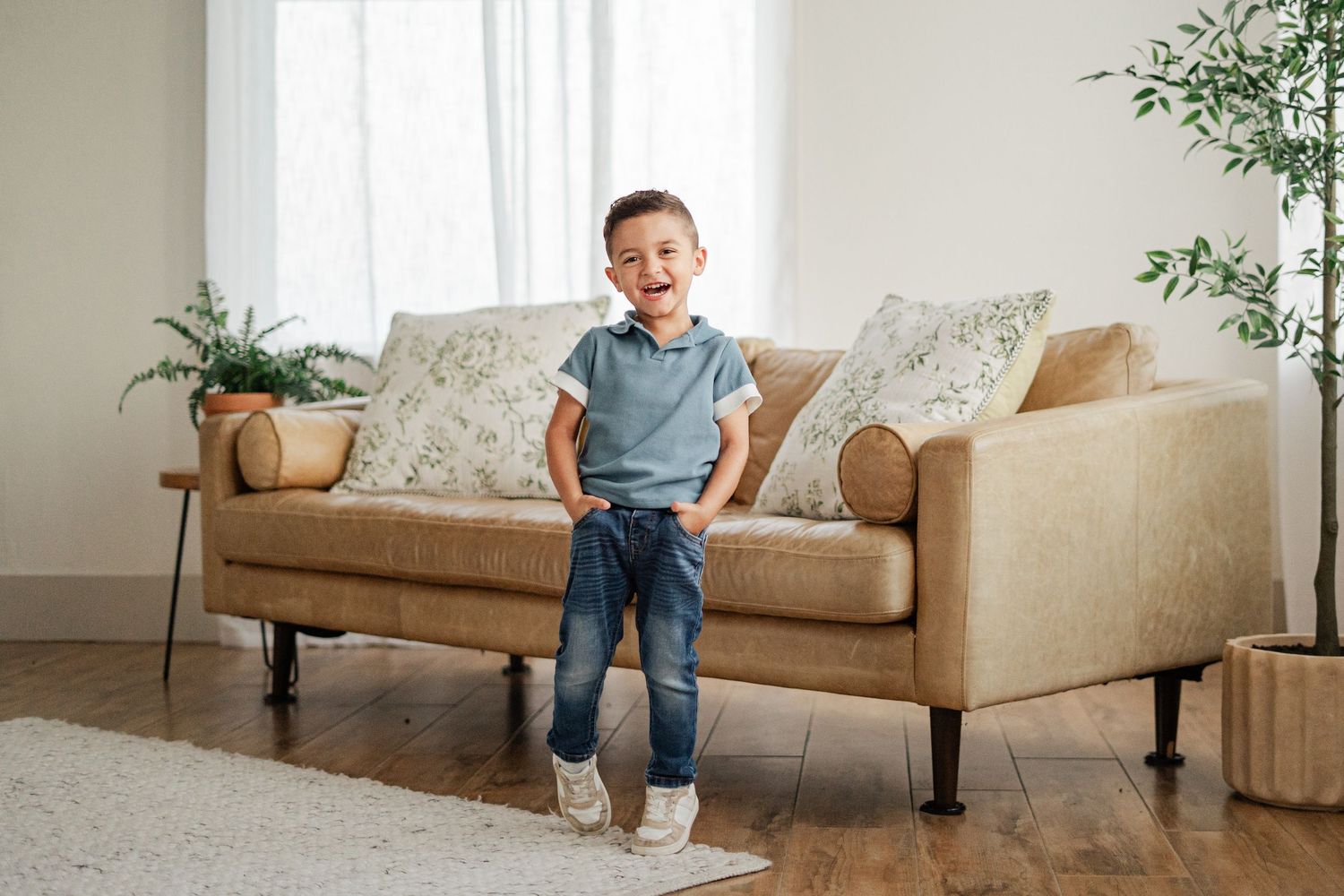 A happy young boy laughing and posing in front of a tan sofa in a bright modern living room.