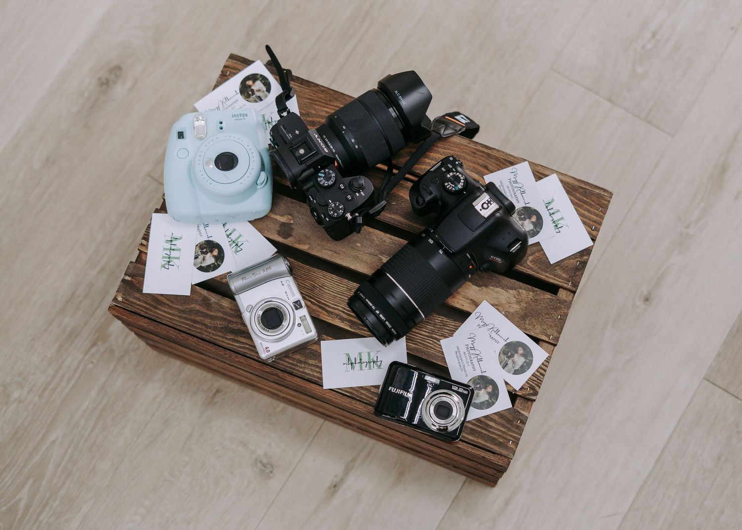 Flat lay of cameras including Fujifilm Instax, DSLRs, and accessories arranged on a rustic wooden crate.