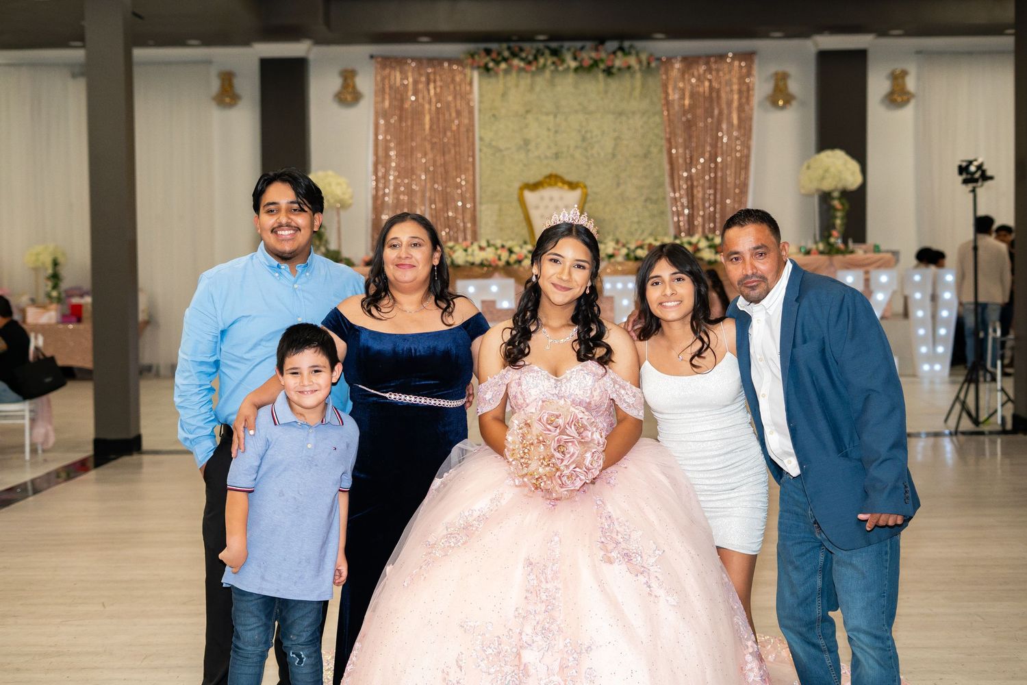 Quinceañera girl in pink gown smiles with six family members in front of decorated celebration backdrop.