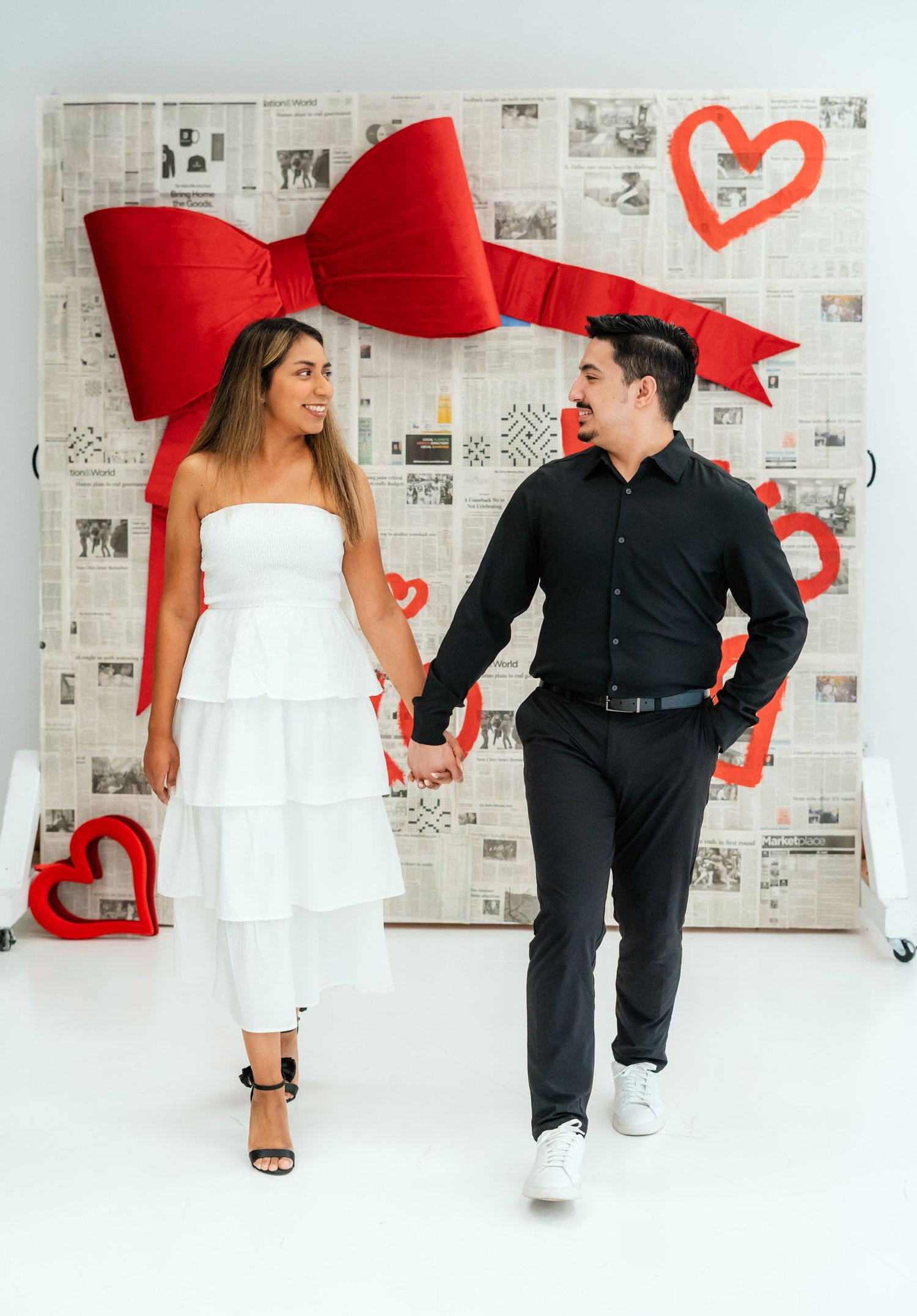 Couple holding hands in front of Valentine's Day newspaper backdrop with red bow and hearts.