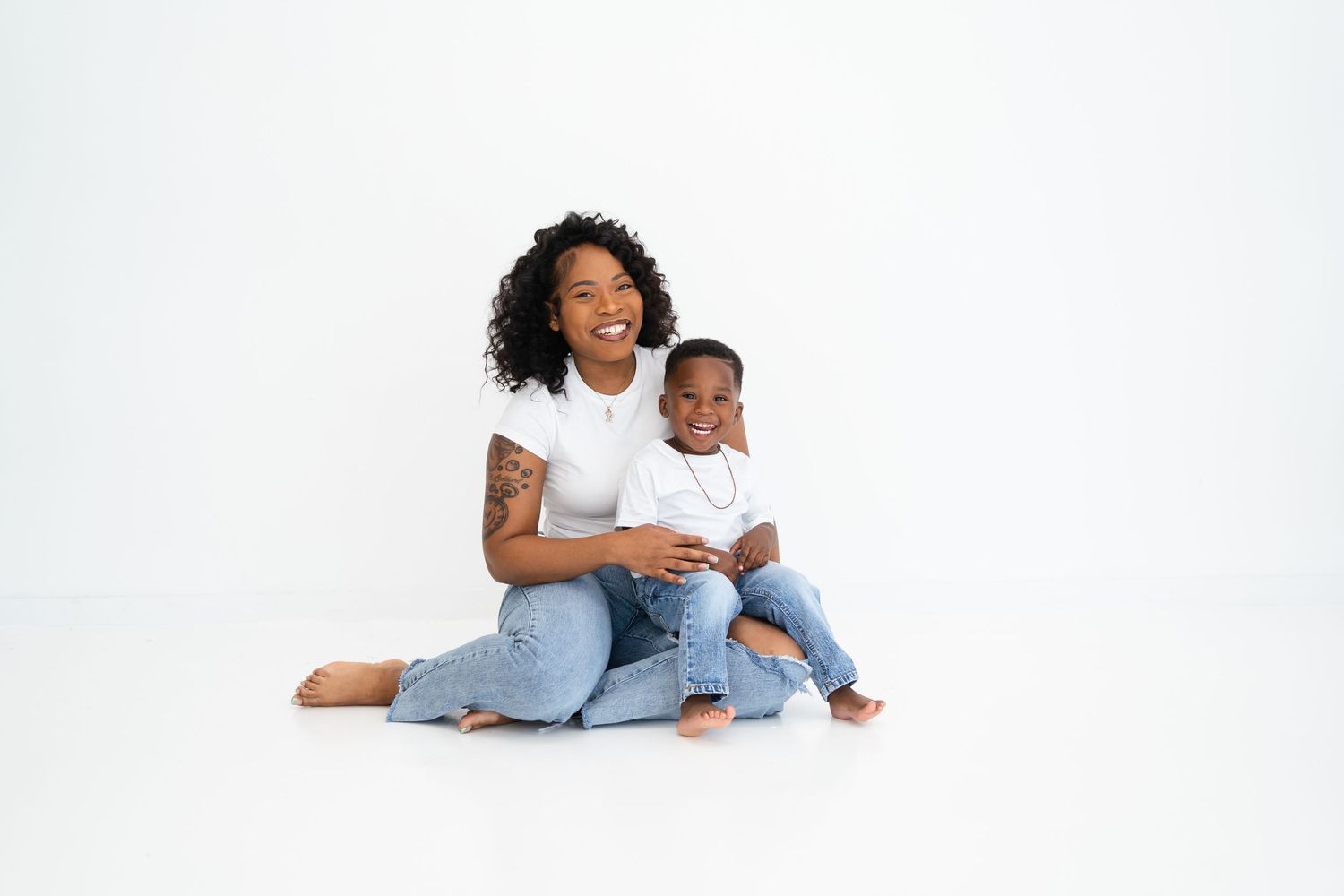 Smiling Black mother hugging her young son in matching white shirts and jeans in white studio.