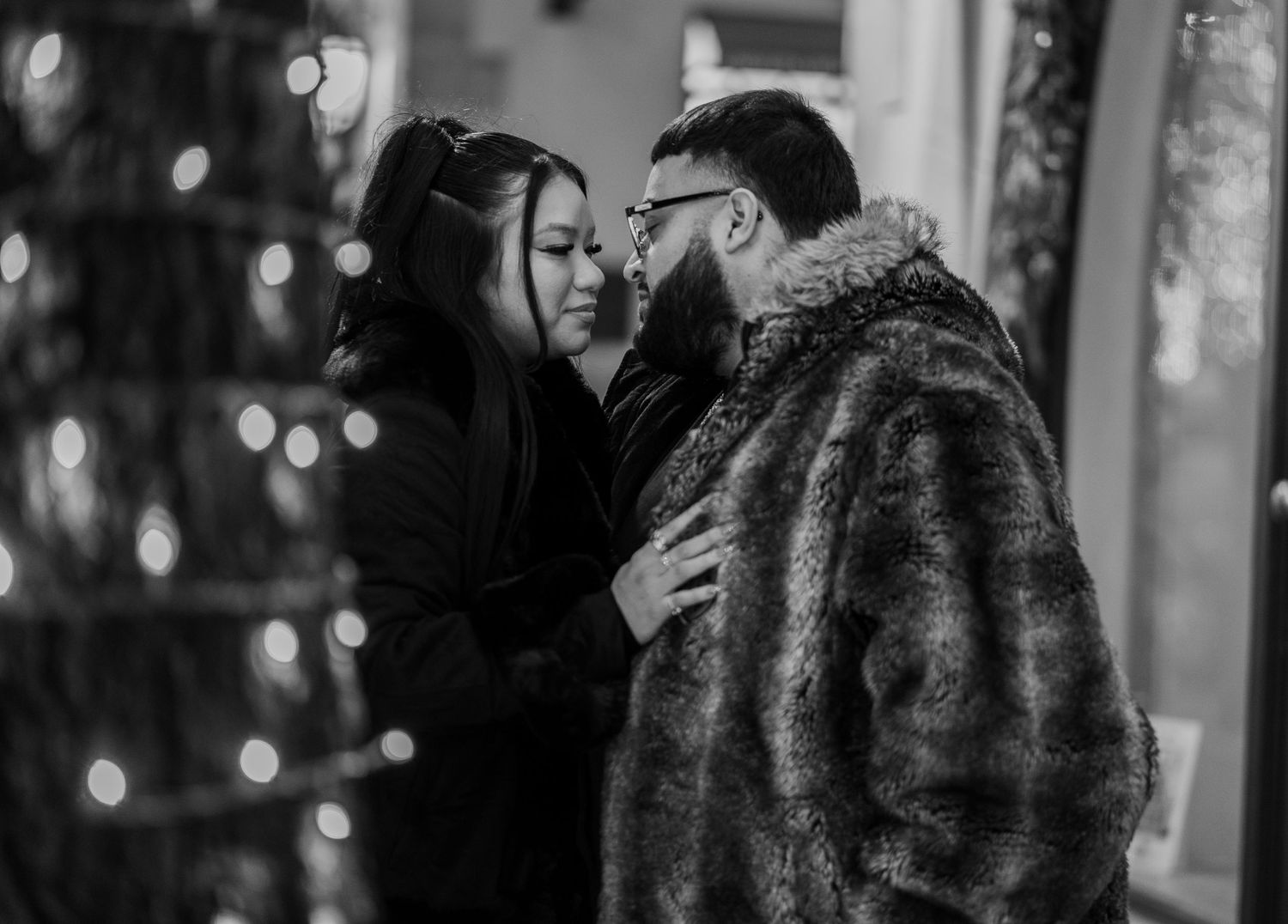 Couple sharing a romantic almost-kiss moment surrounded by bokeh holiday lights, black and white photo.