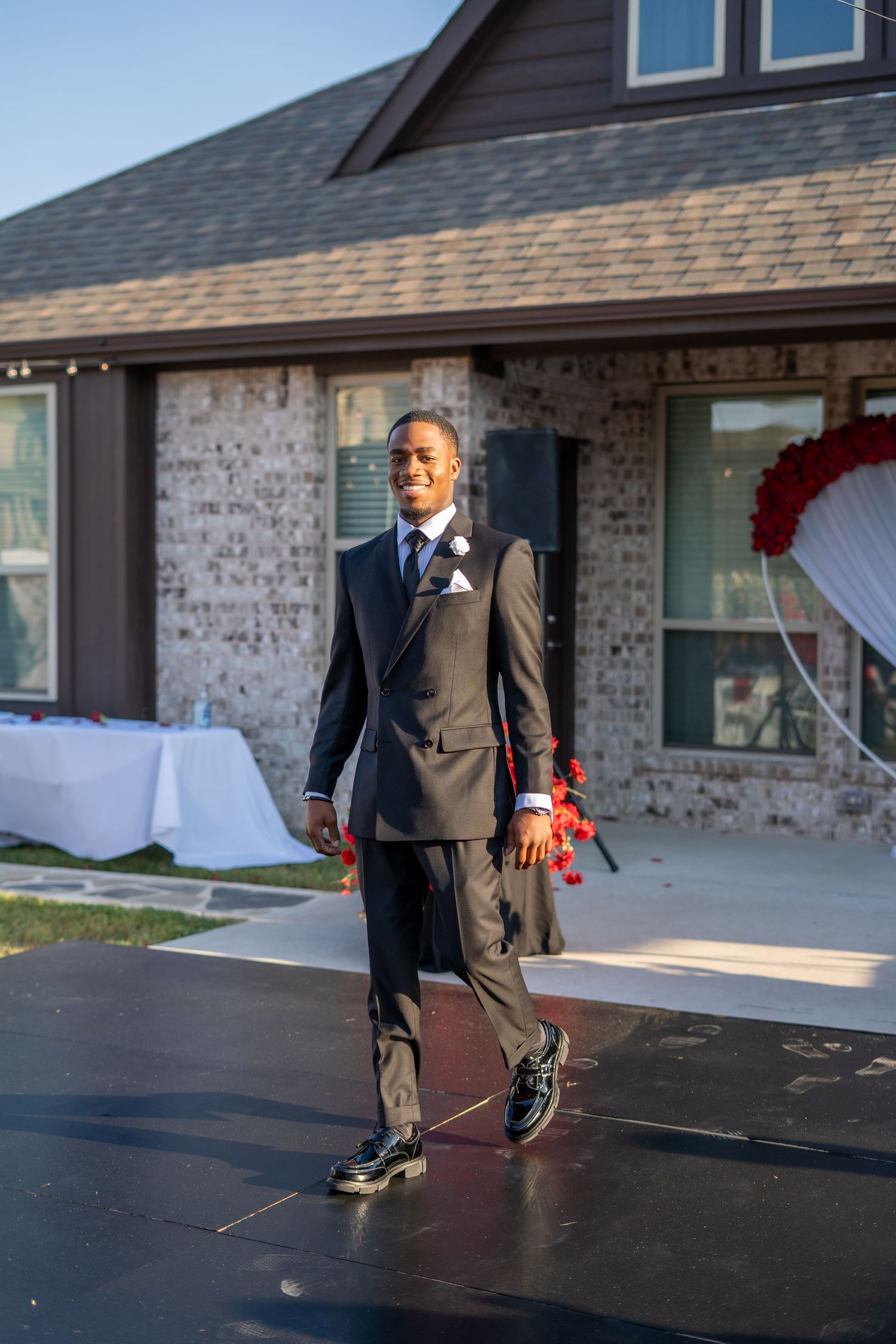 Dapper groom in black suit walks confidently on outdoor stage at wedding venue decorated with red roses.