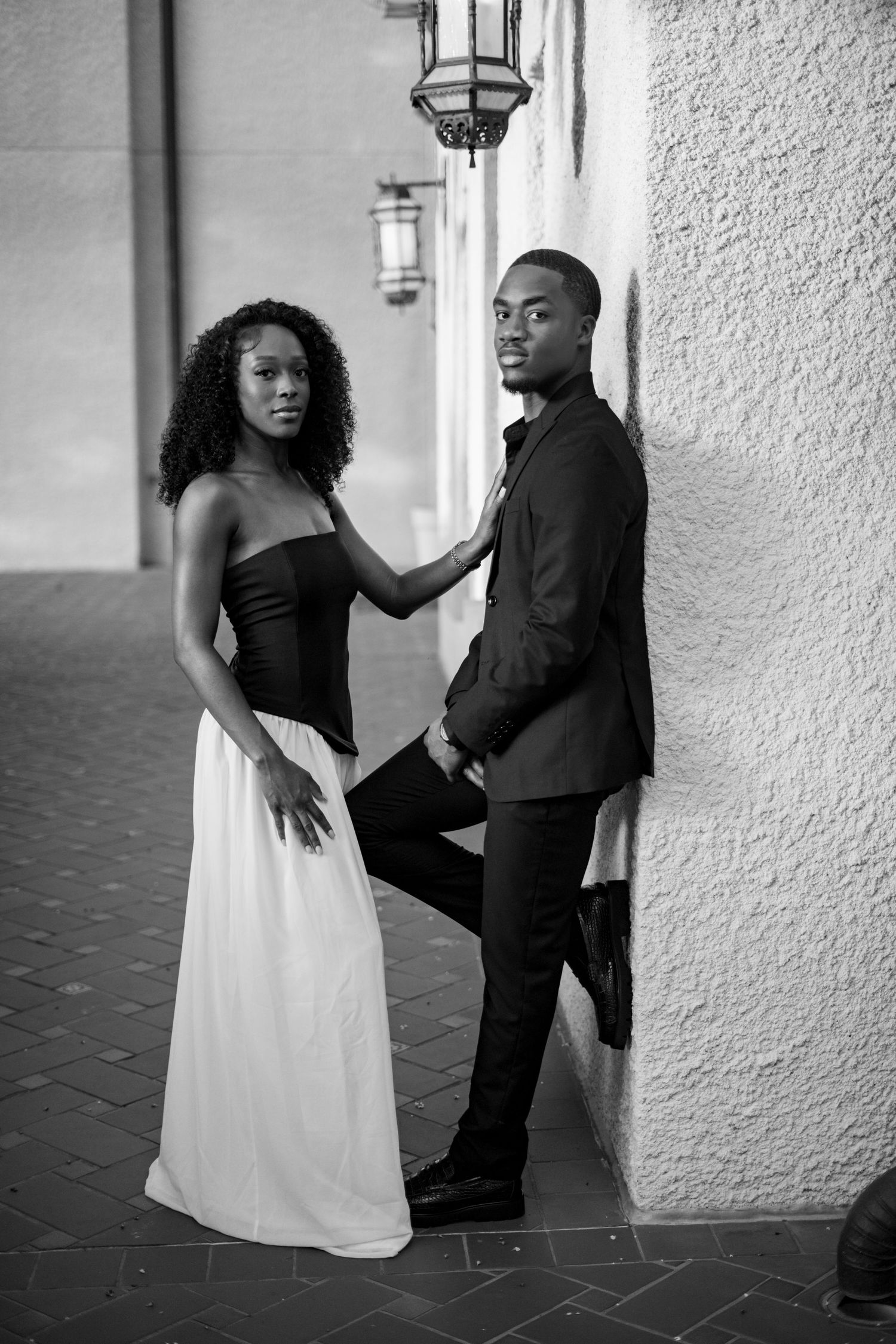 Black and white portrait of a stylish couple posing against a textured wall under a vintage lantern.