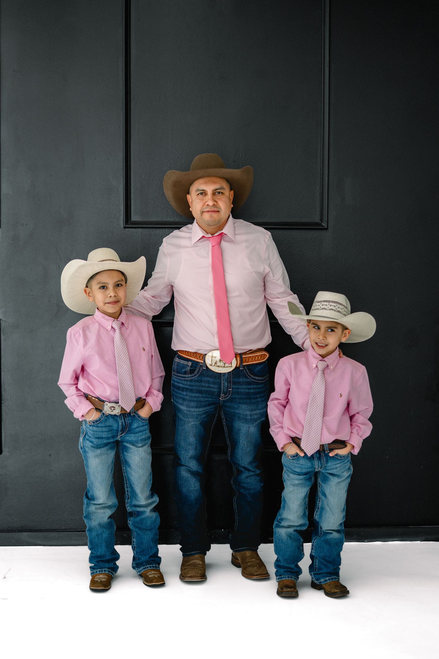 A father and two young boys in matching pink shirts, ties, jeans, and cowboy hats pose for a family portrait.