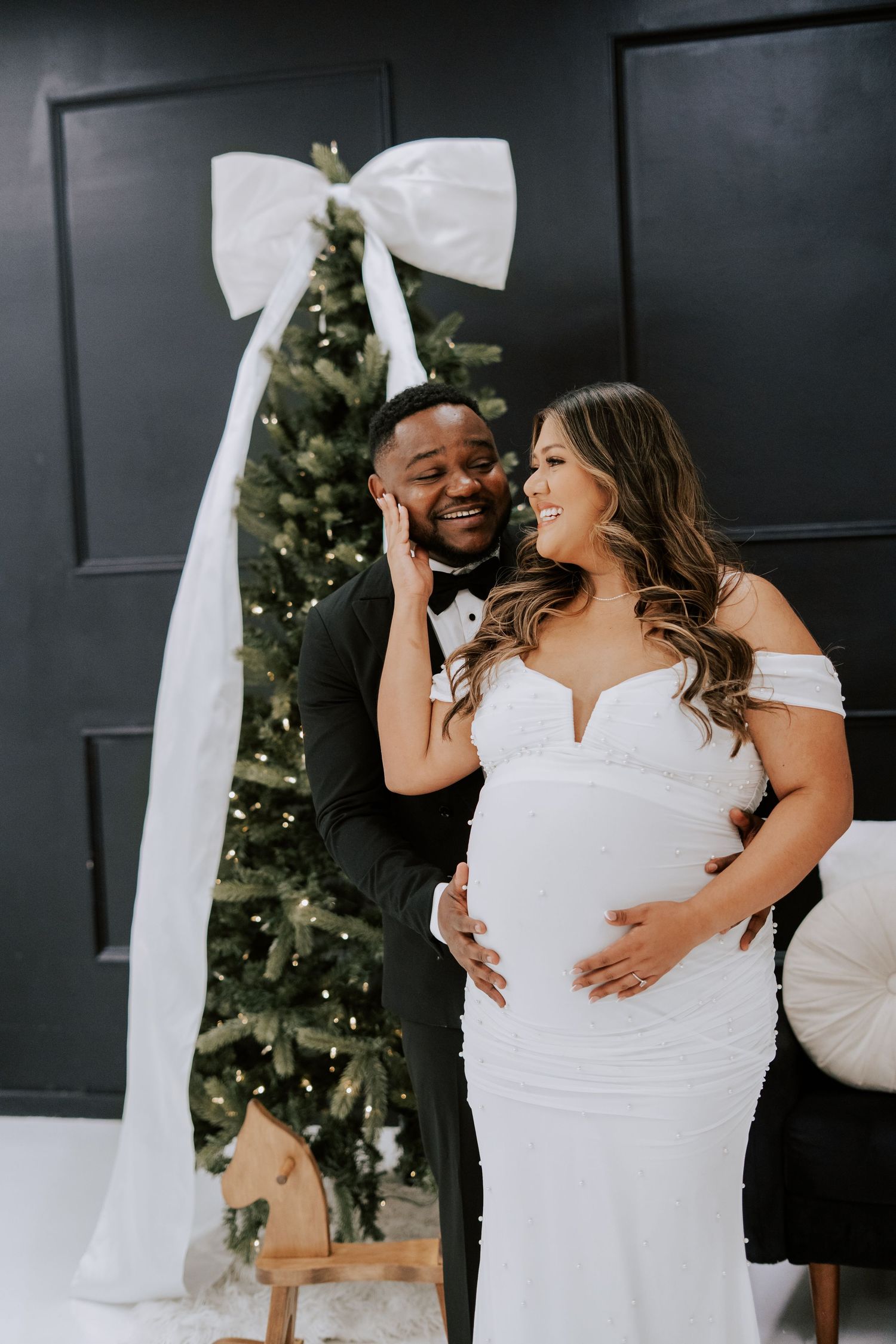 Expecting couple smiling together in front of decorated Christmas tree with large white bow backdrop.