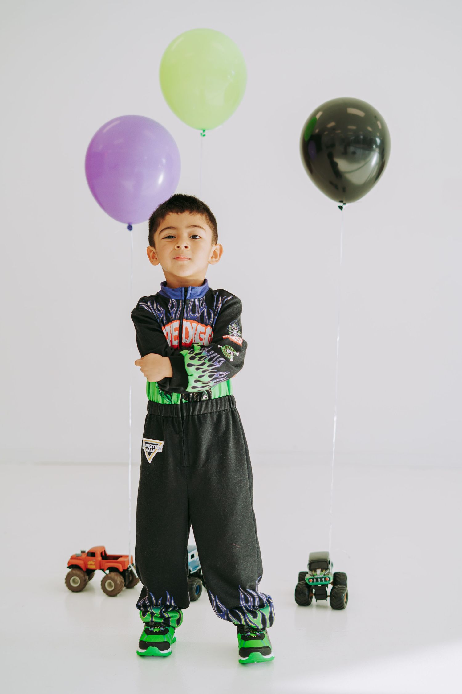 Young boy in Monster Jam racing suit poses with arms crossed beside balloons and monster truck toys.