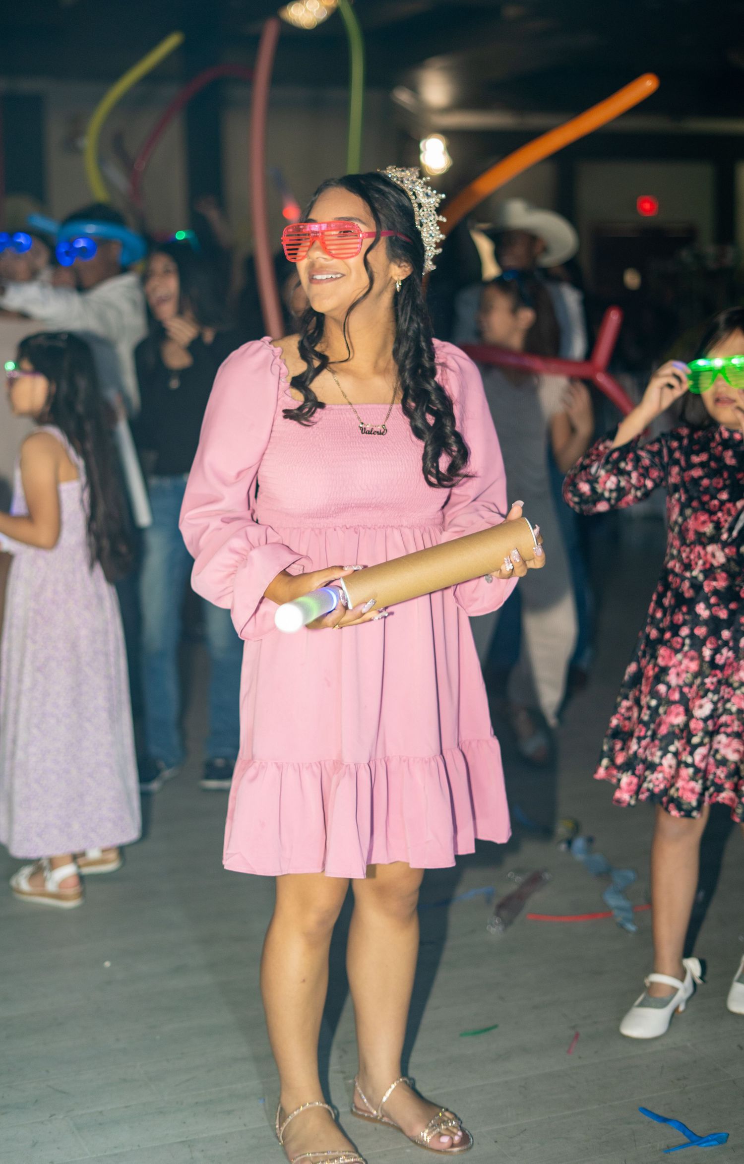 Quinceañera girl in pink dress and tiara dances with heart-shaped glasses and confetti cannon.