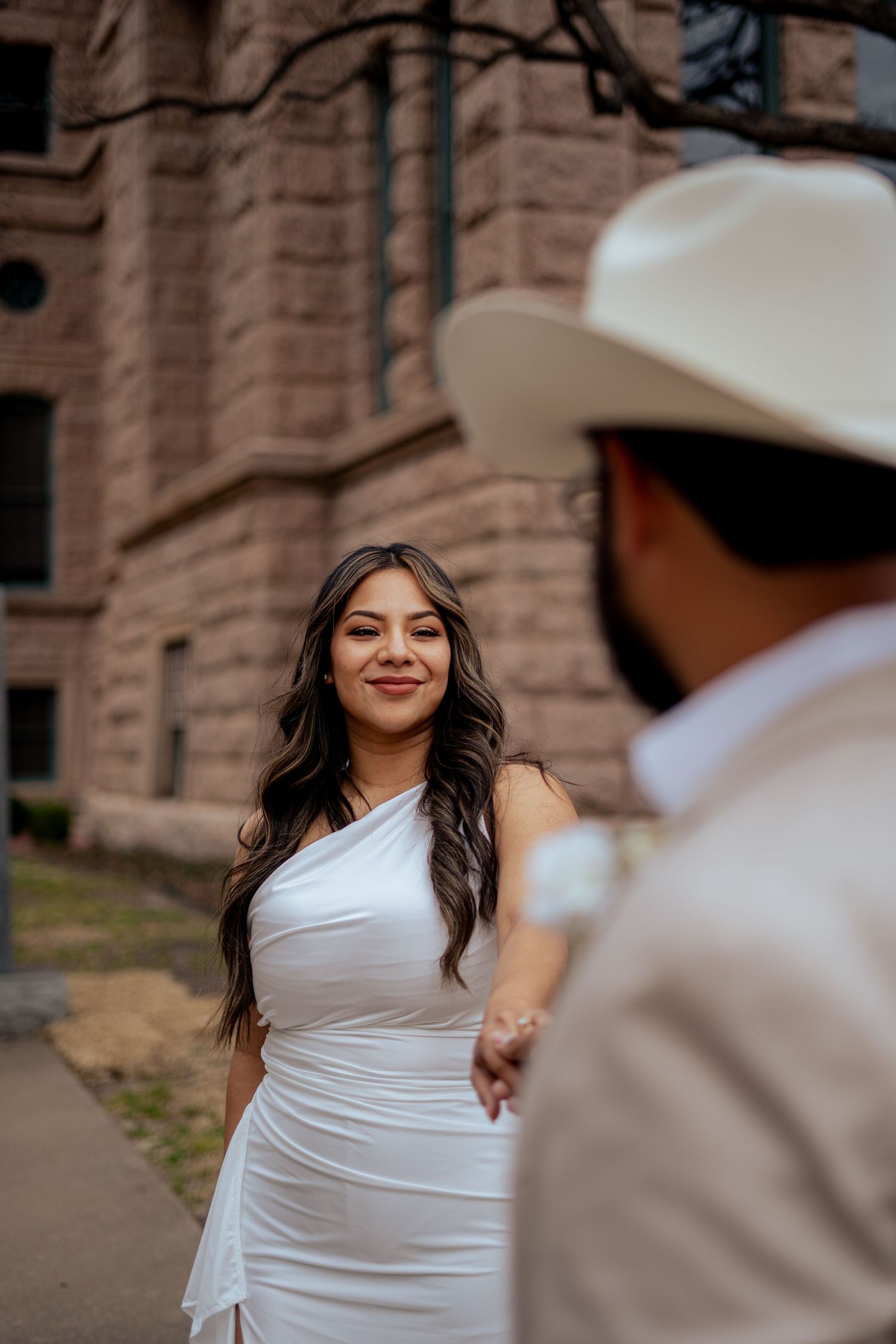 Smiling bride in white one-shoulder gown holds groom's hand outside historic courthouse building.