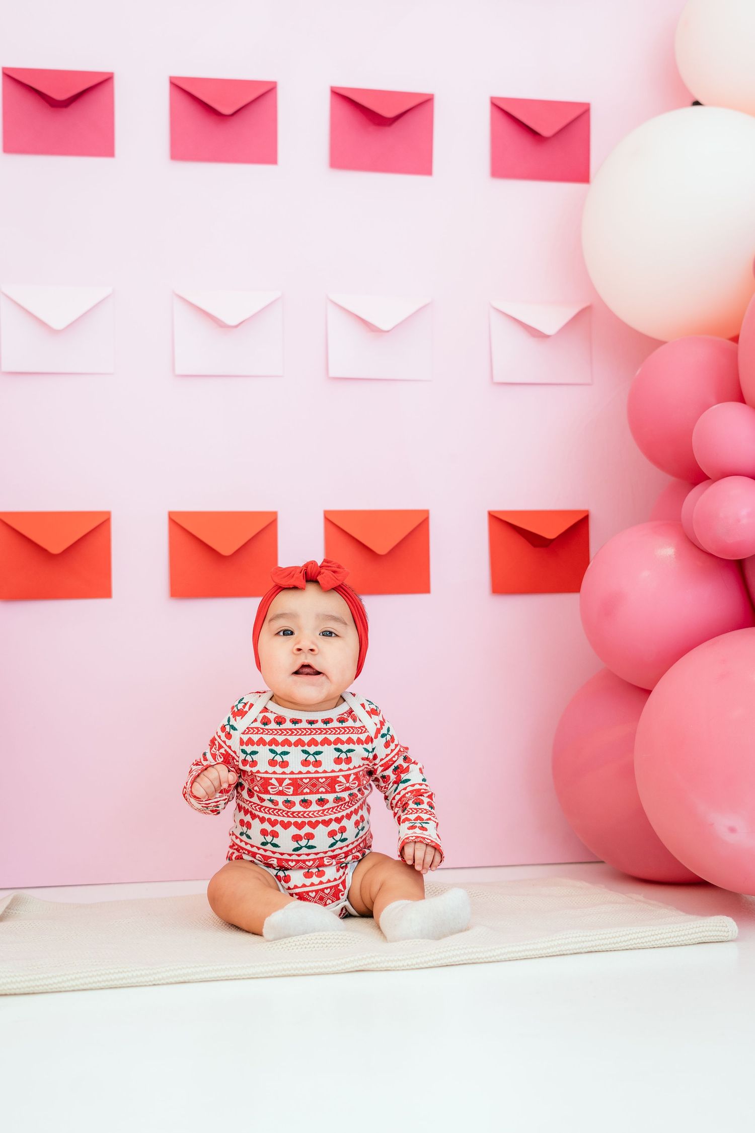 Baby in festive onesie sits before pink wall with envelopes and balloons.