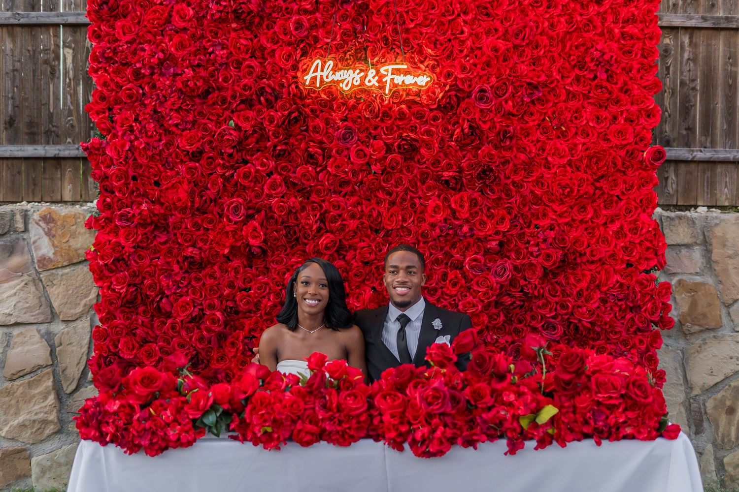 Bride and groom sit together at sweetheart table surrounded by red roses beneath neon sign.