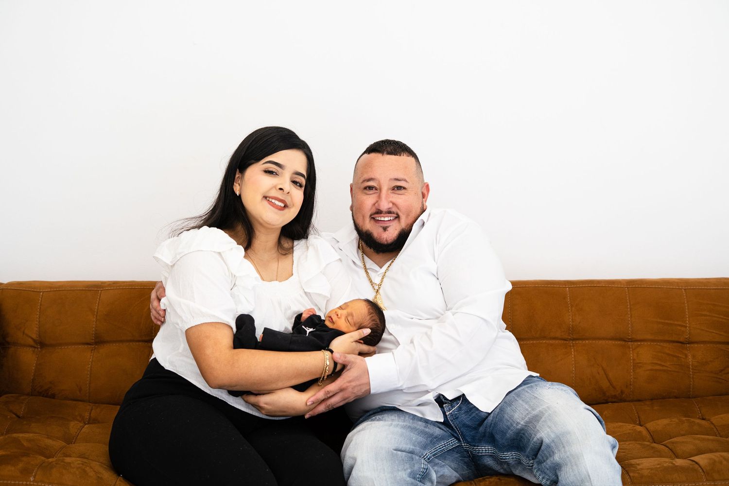 A couple smiles warmly while cradling their newborn baby on a brown leather couch.
