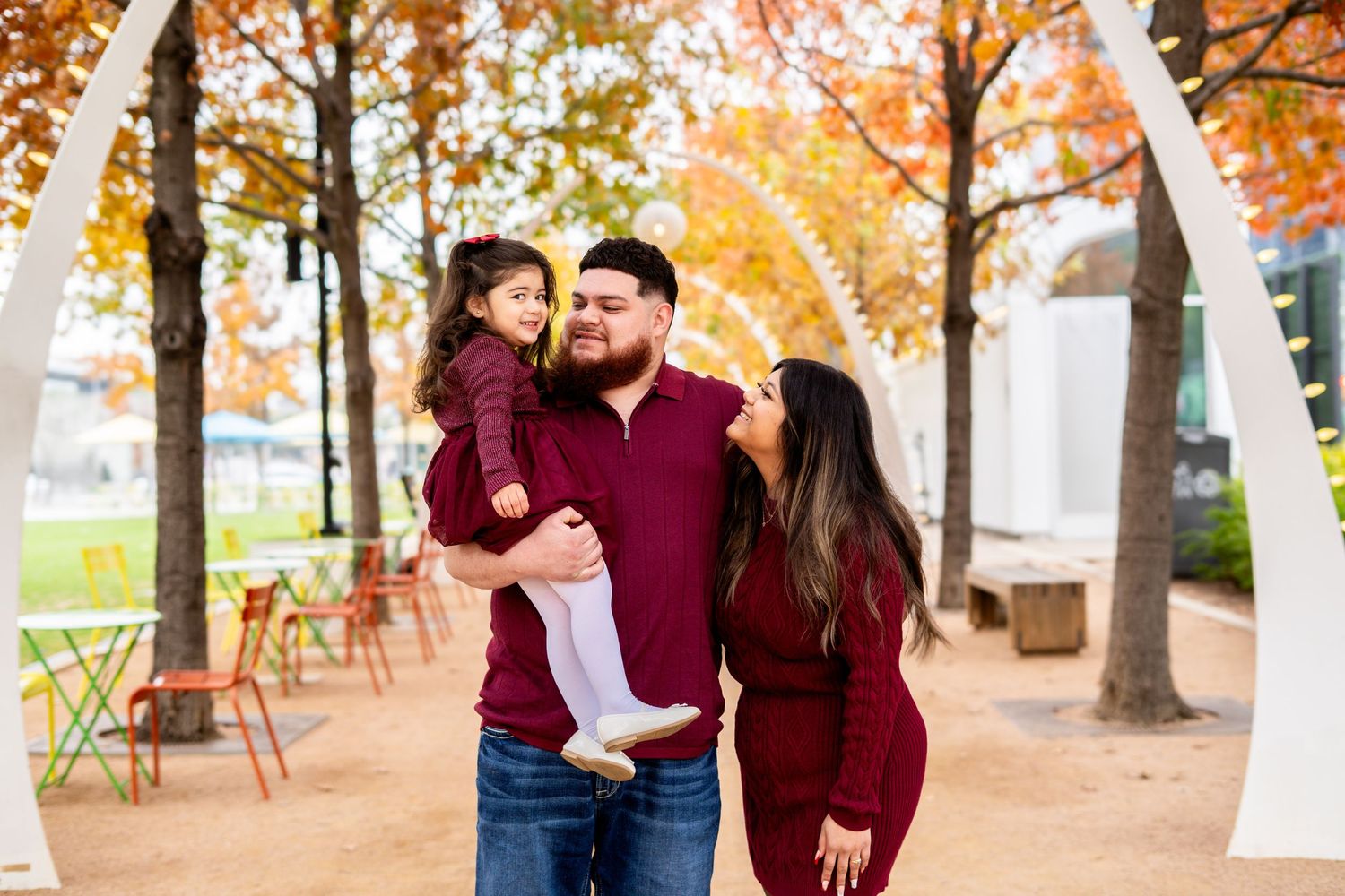 Father holding young daughter while mother stands close, smiling together under fall foliage.
