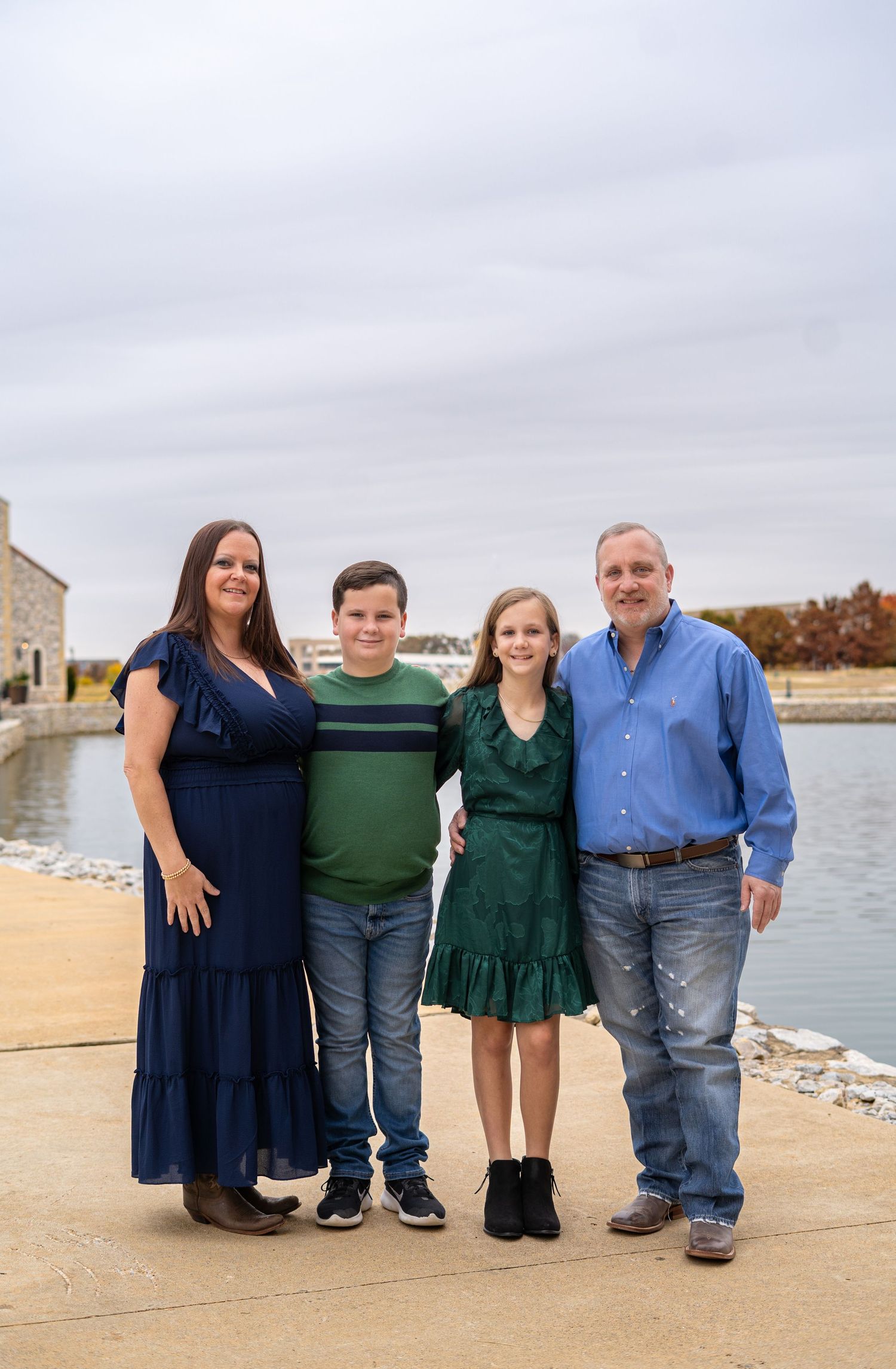 Family of four posing by a waterfront on an overcast autumn day in coordinating blue and green outfits.