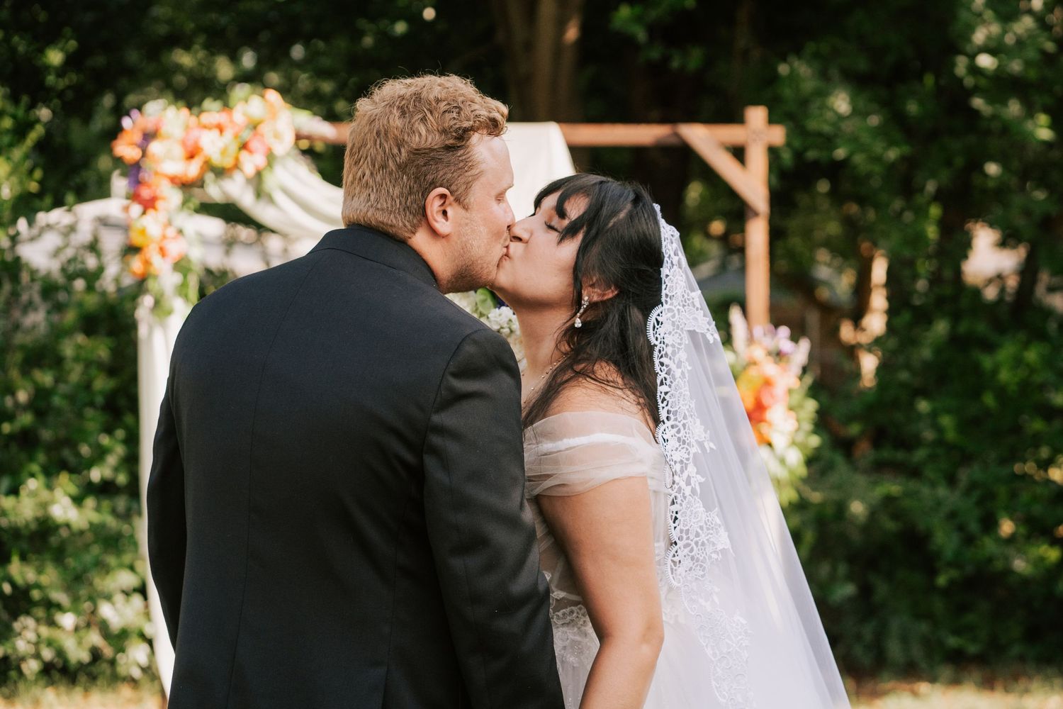 Bride and groom share first kiss at outdoor wedding ceremony with floral arch backdrop.