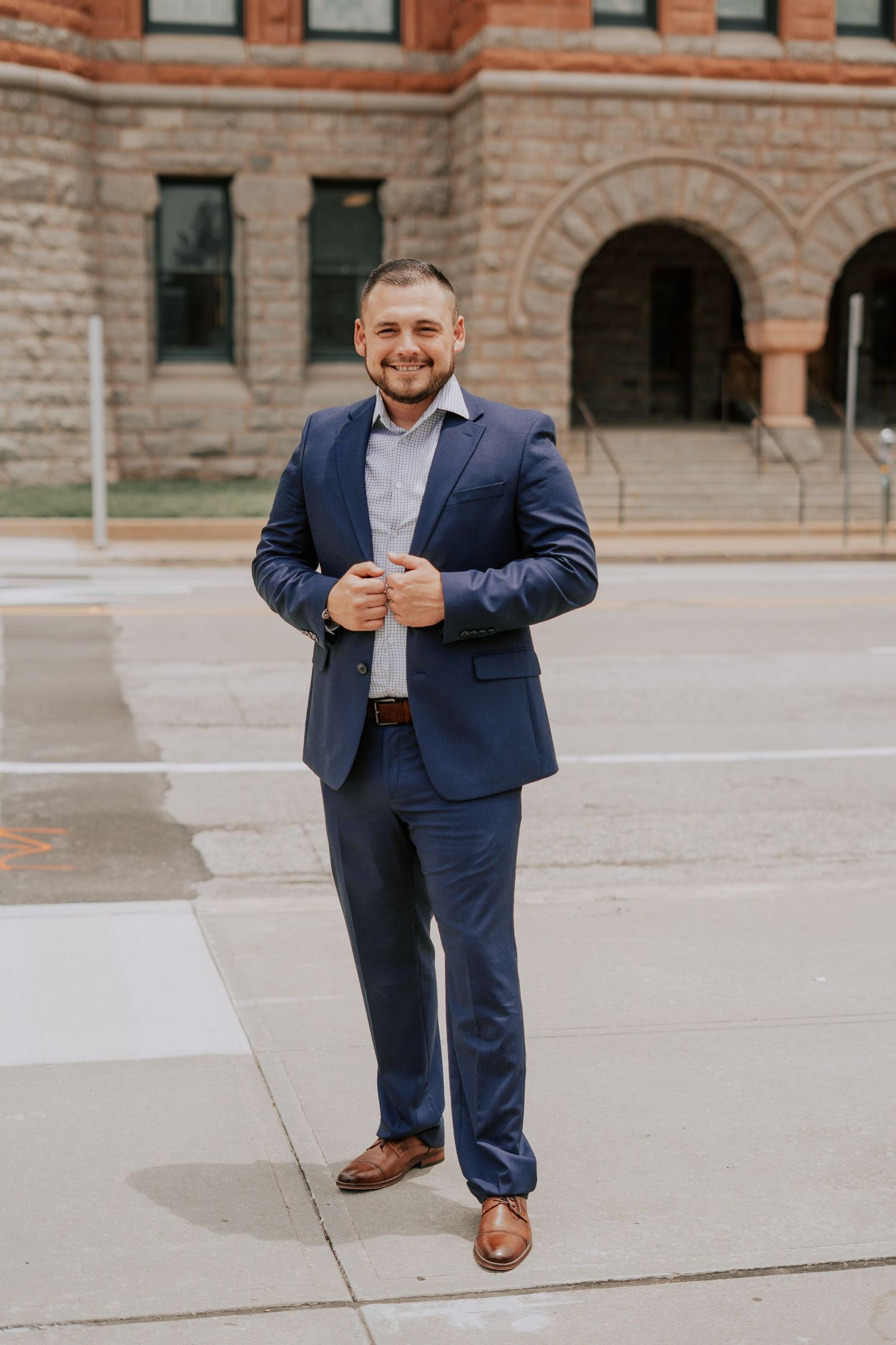 Smiling businessman in navy suit standing on sidewalk in front of historic red brick building.