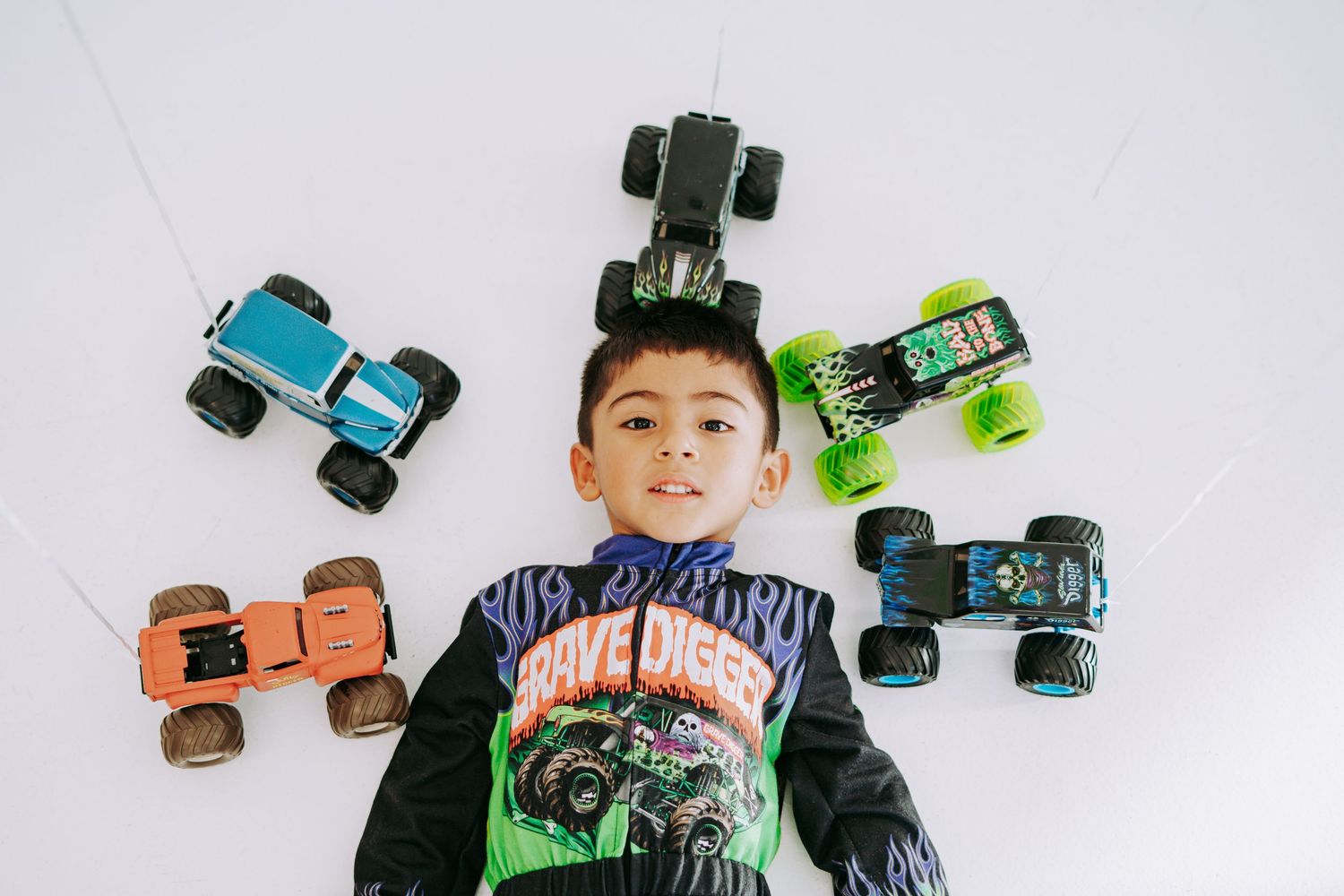 Boy lying on floor surrounded by four monster truck toys in aerial flat lay photo.