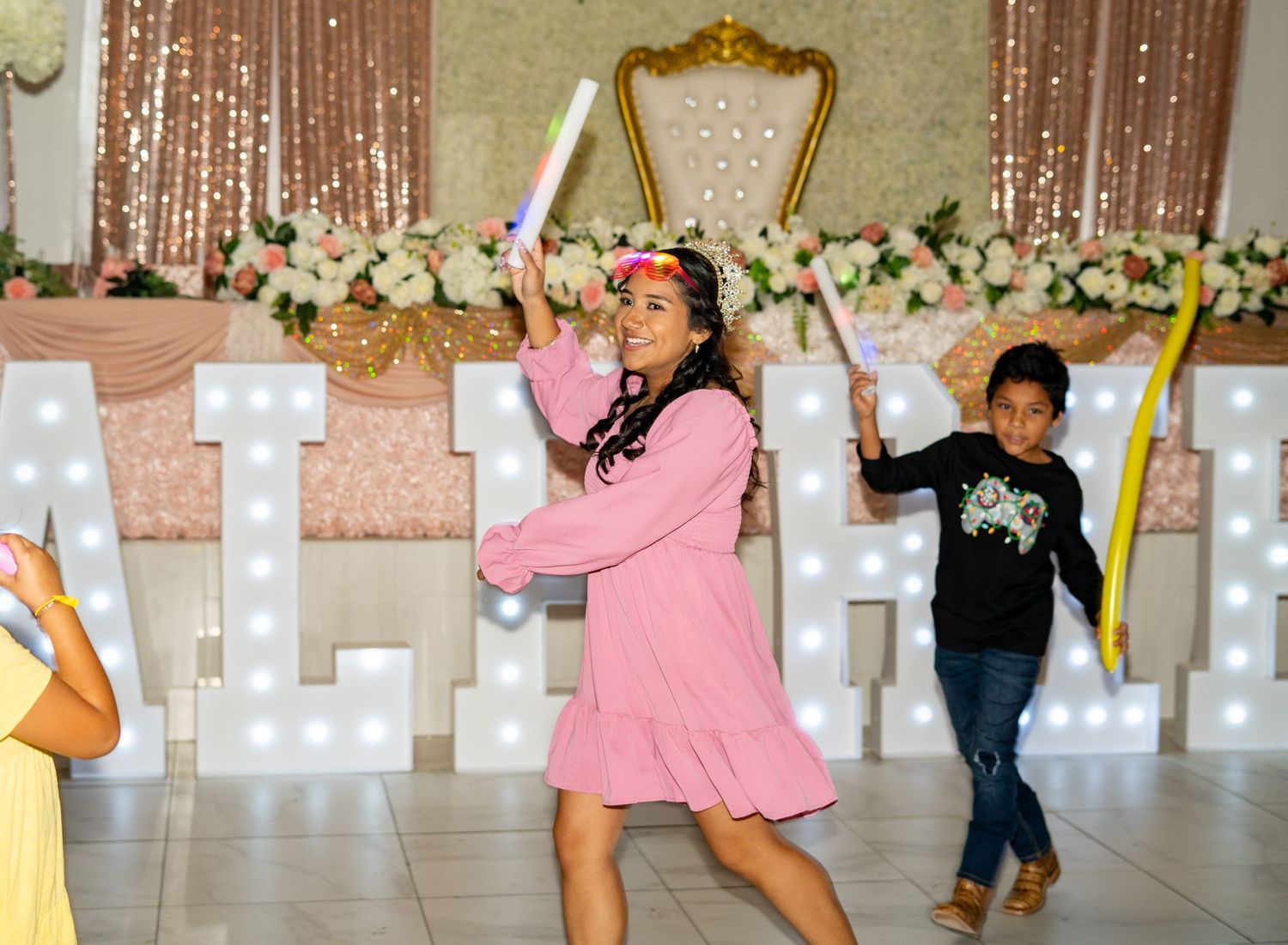 Girl in pink dress dances joyfully in front of illuminated letter name display at quinceañera.