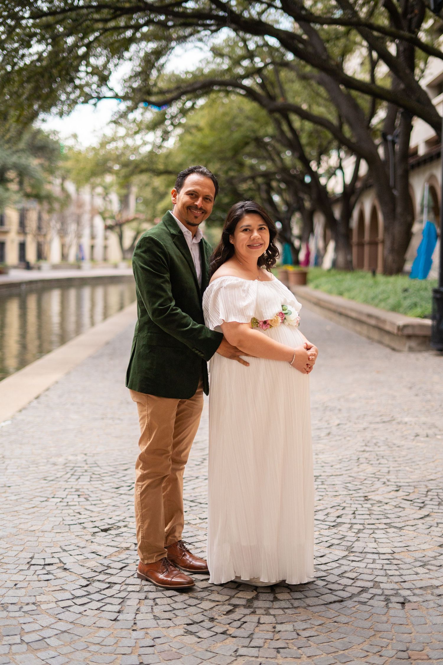 Couple posing on cobblestone path, man in green blazer embracing pregnant woman in white off-shoulder gown.