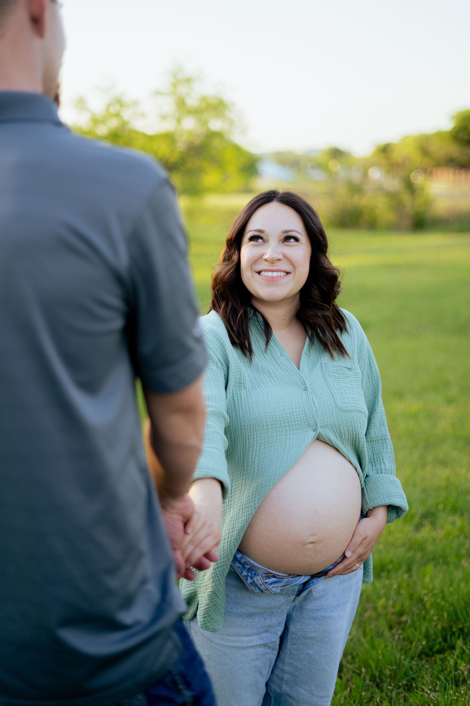 Glowing pregnant woman in mint shirt holds baby bump while smiling at partner during outdoor maternity photoshoot.