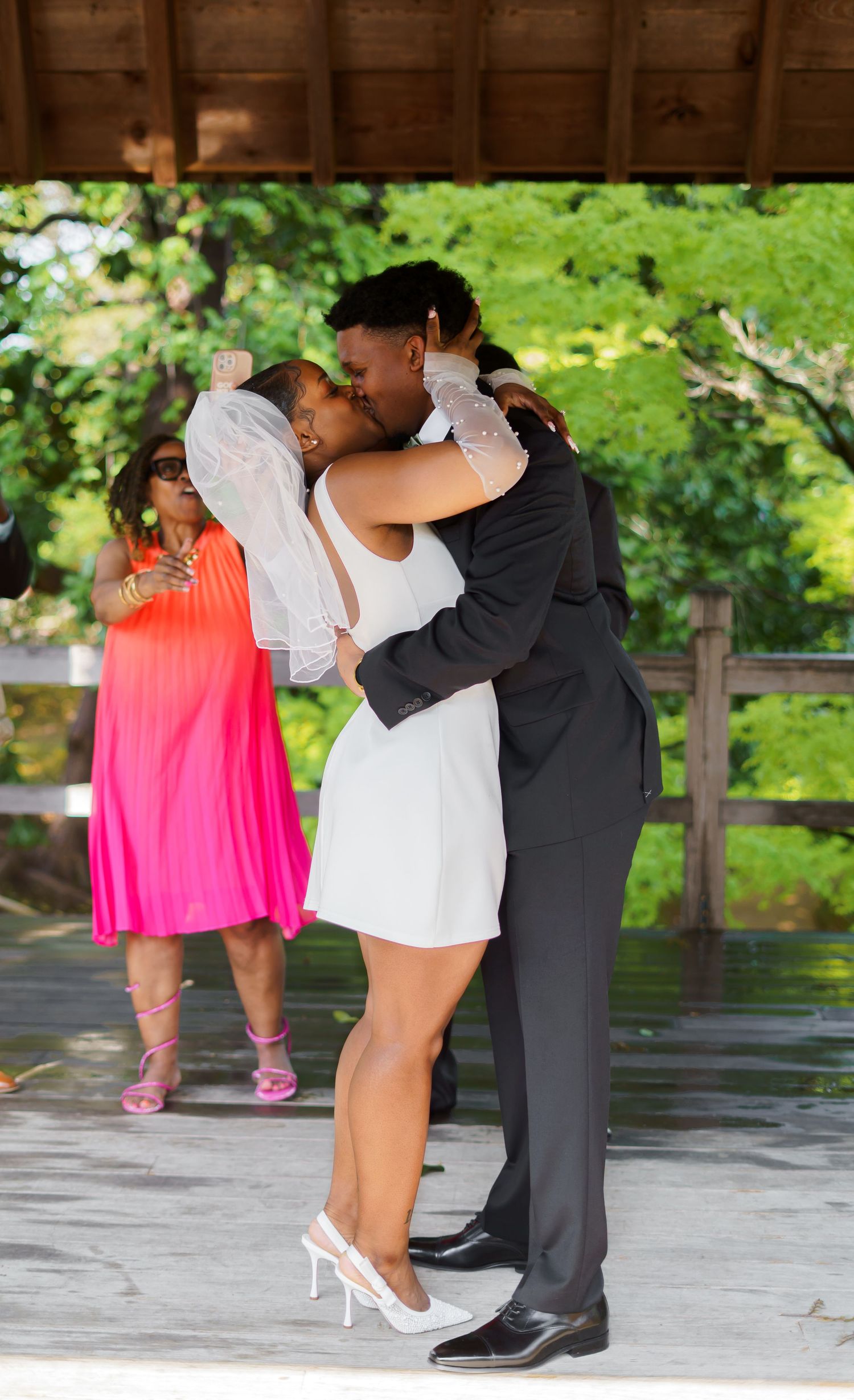 Newlyweds share their first kiss at outdoor wedding ceremony as guest in pink dress watches nearby.