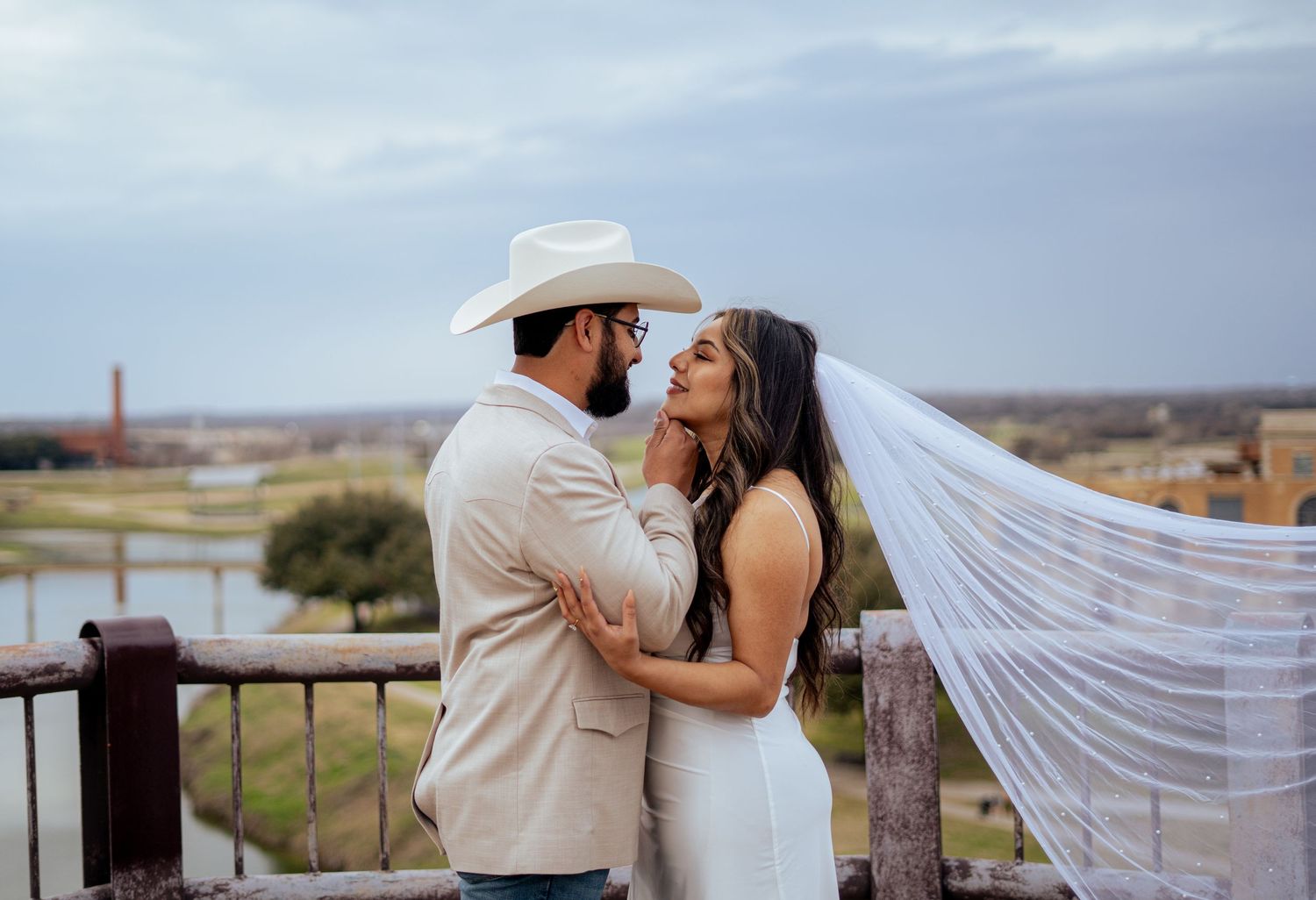 Couple in wedding attire sharing a romantic moment on a deck, groom in cowboy hat, bride's veil flowing in the wind.