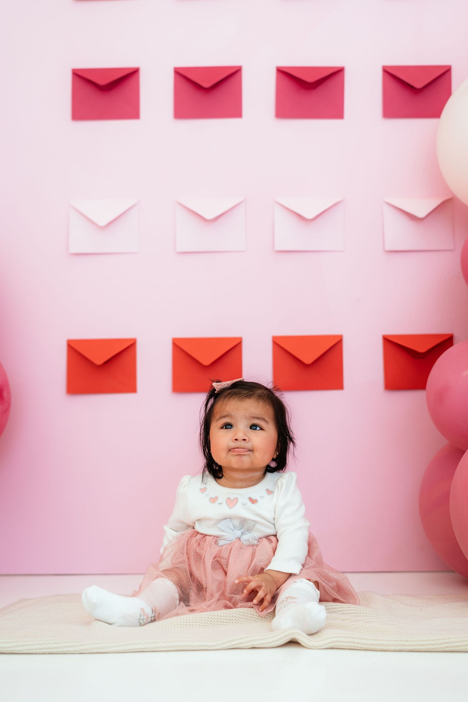 Toddler in white top and pink tutu sits before pink Valentine envelope wall.