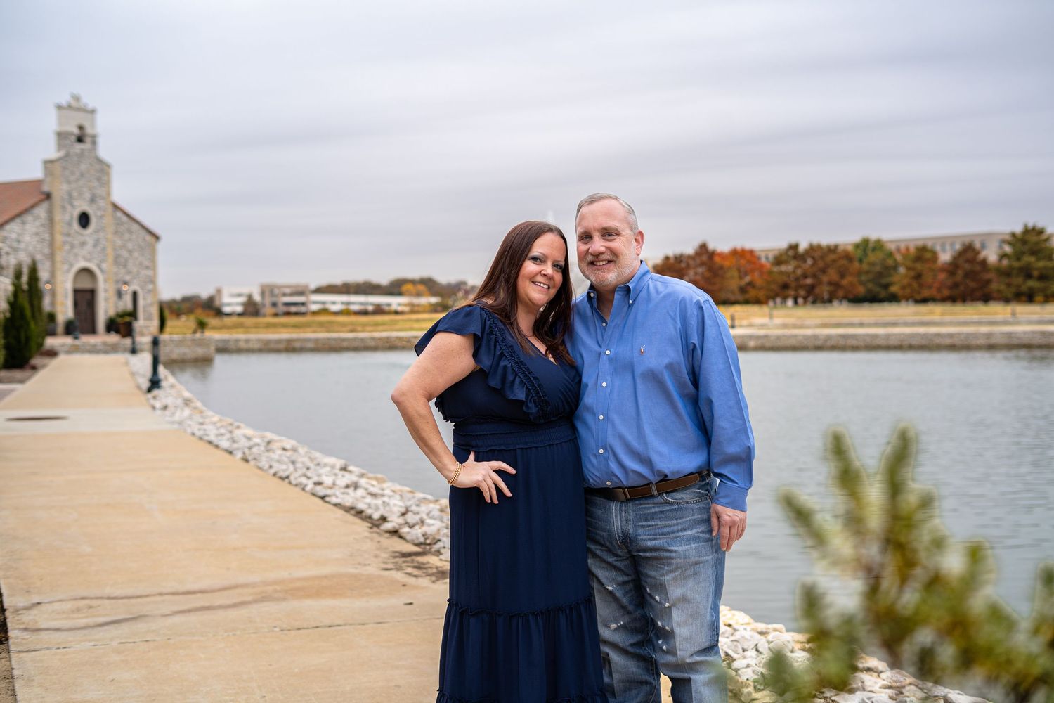 Couple smiling together by a rocky waterfront with a stone chapel in the background on a cloudy day.