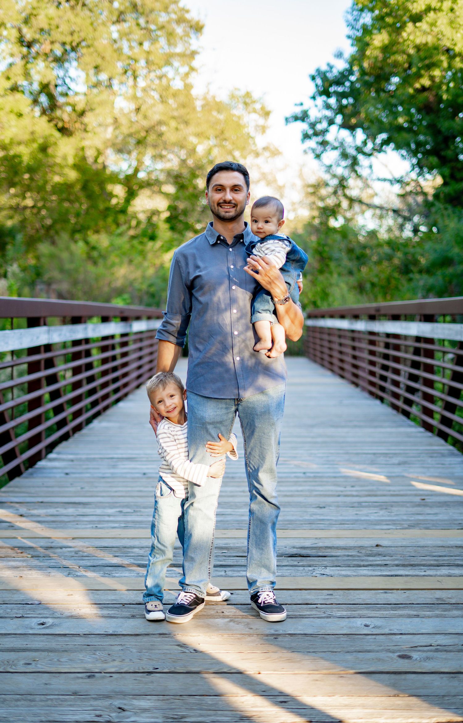 Smiling father holding two young children on a wooden bridge surrounded by green trees.