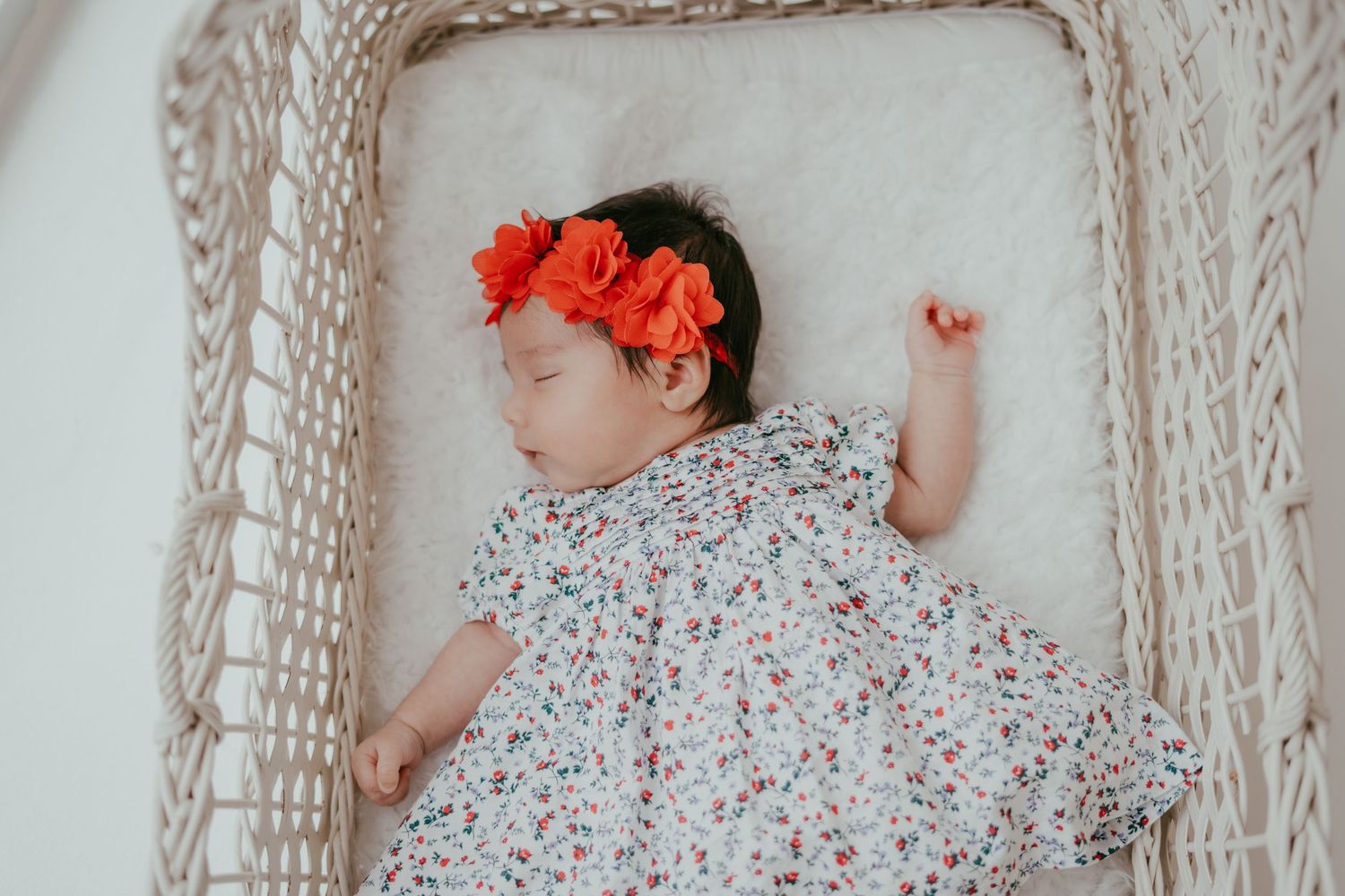 Sleeping newborn baby girl in floral outfit with large red flower headband rests in a wicker bassinet.