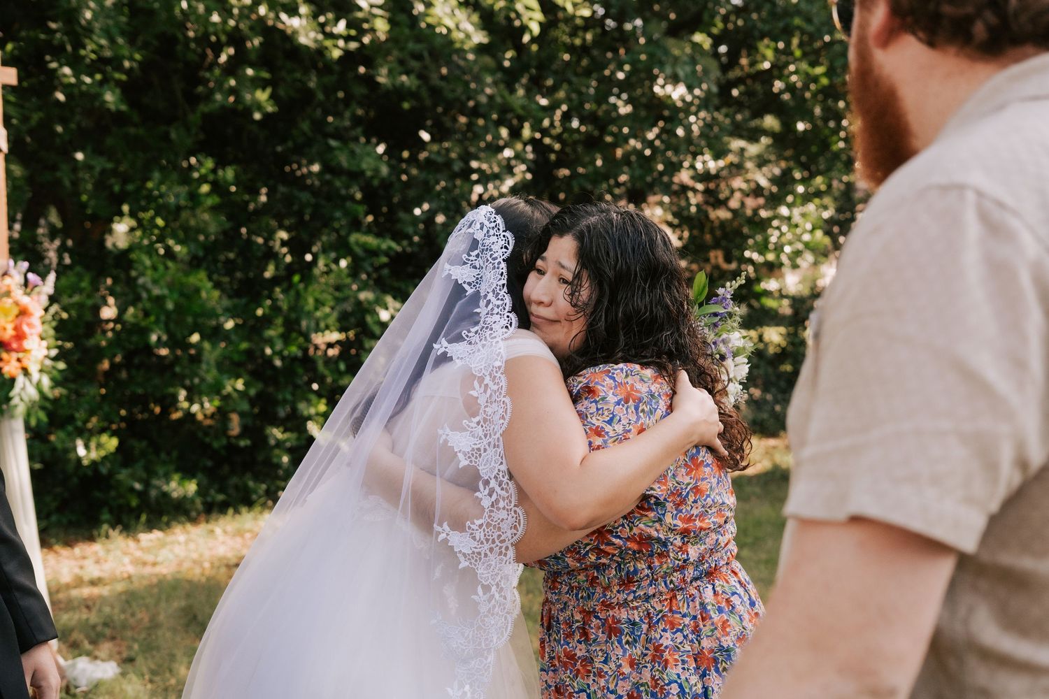 Bride in lace veil embracing a guest in floral dress at outdoor wedding ceremony.