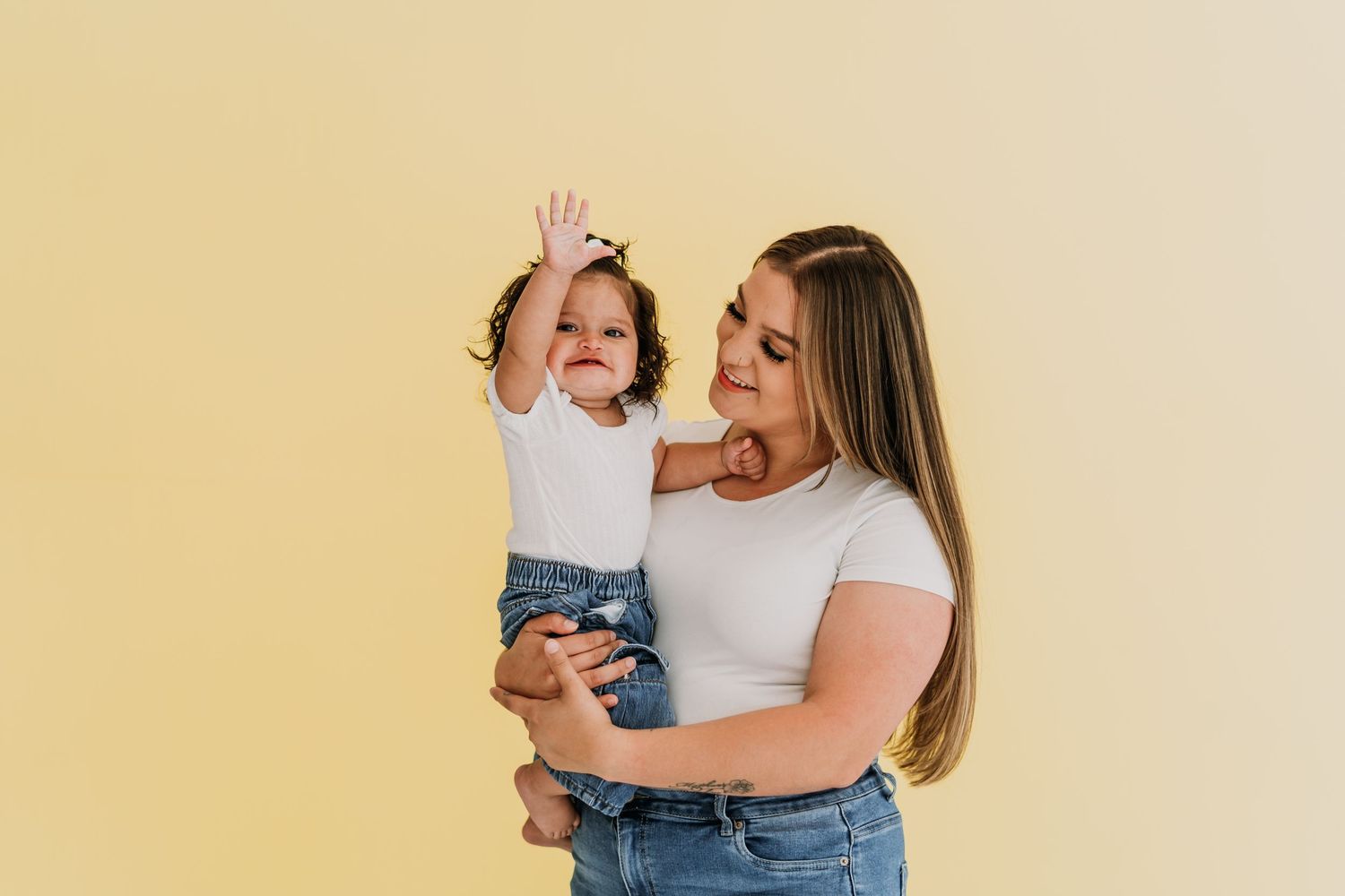 A young woman in a white top holds a happy baby girl in denim overalls against a soft yellow background.