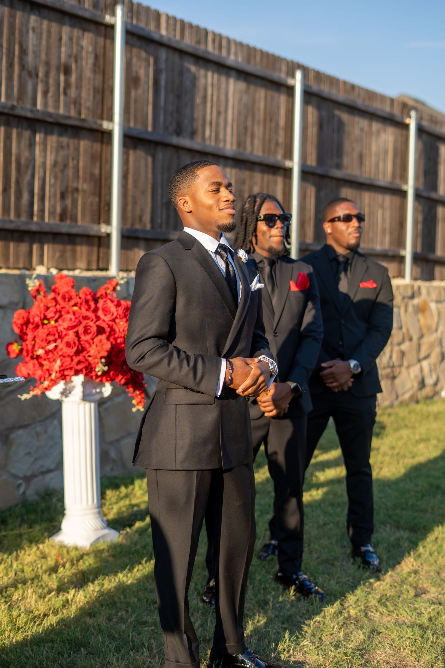 Groom and groomsmen in black suits stand outdoors near red floral arrangements at wedding ceremony.