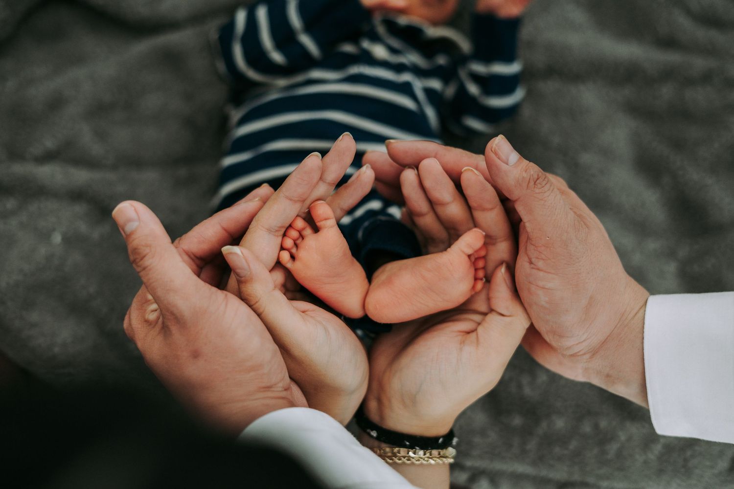 Parents gently cupping newborn baby's tiny feet together in their hands on gray blanket.