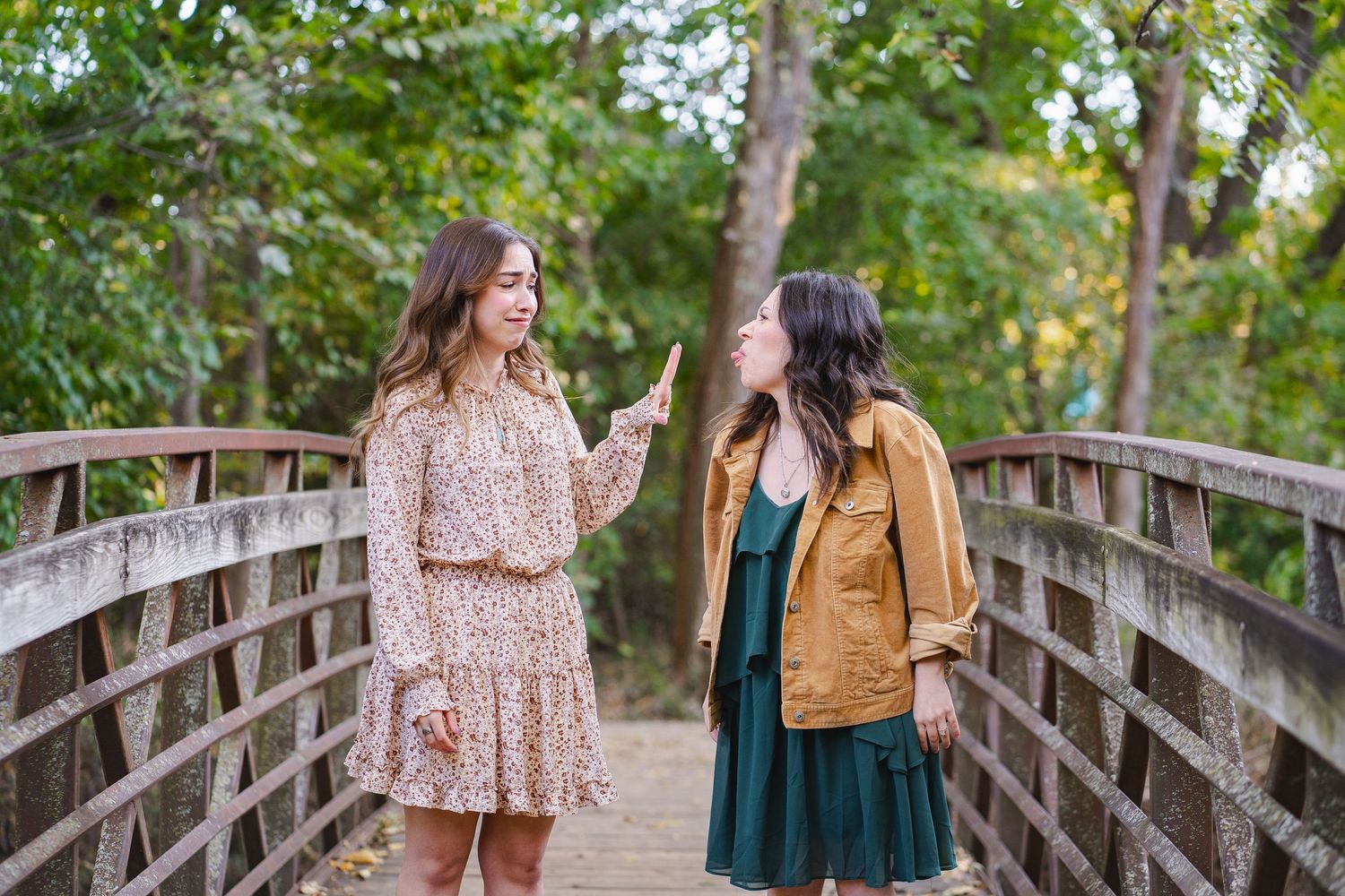 Two young women laughing and talking together on a wooden bridge surrounded by green trees.