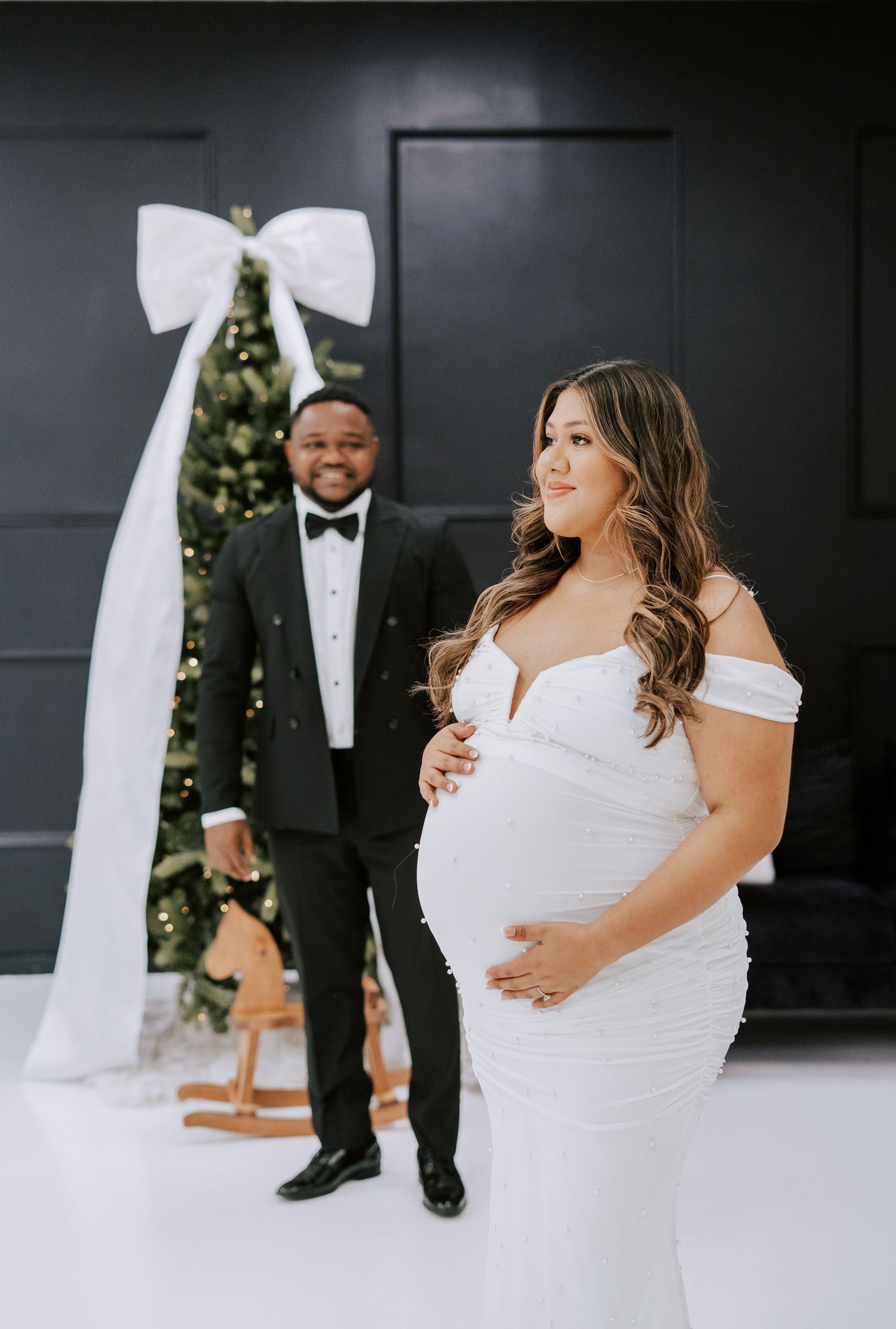 Maternity photo of pregnant woman in white gown with partner in tuxedo before Christmas tree with white bow.
